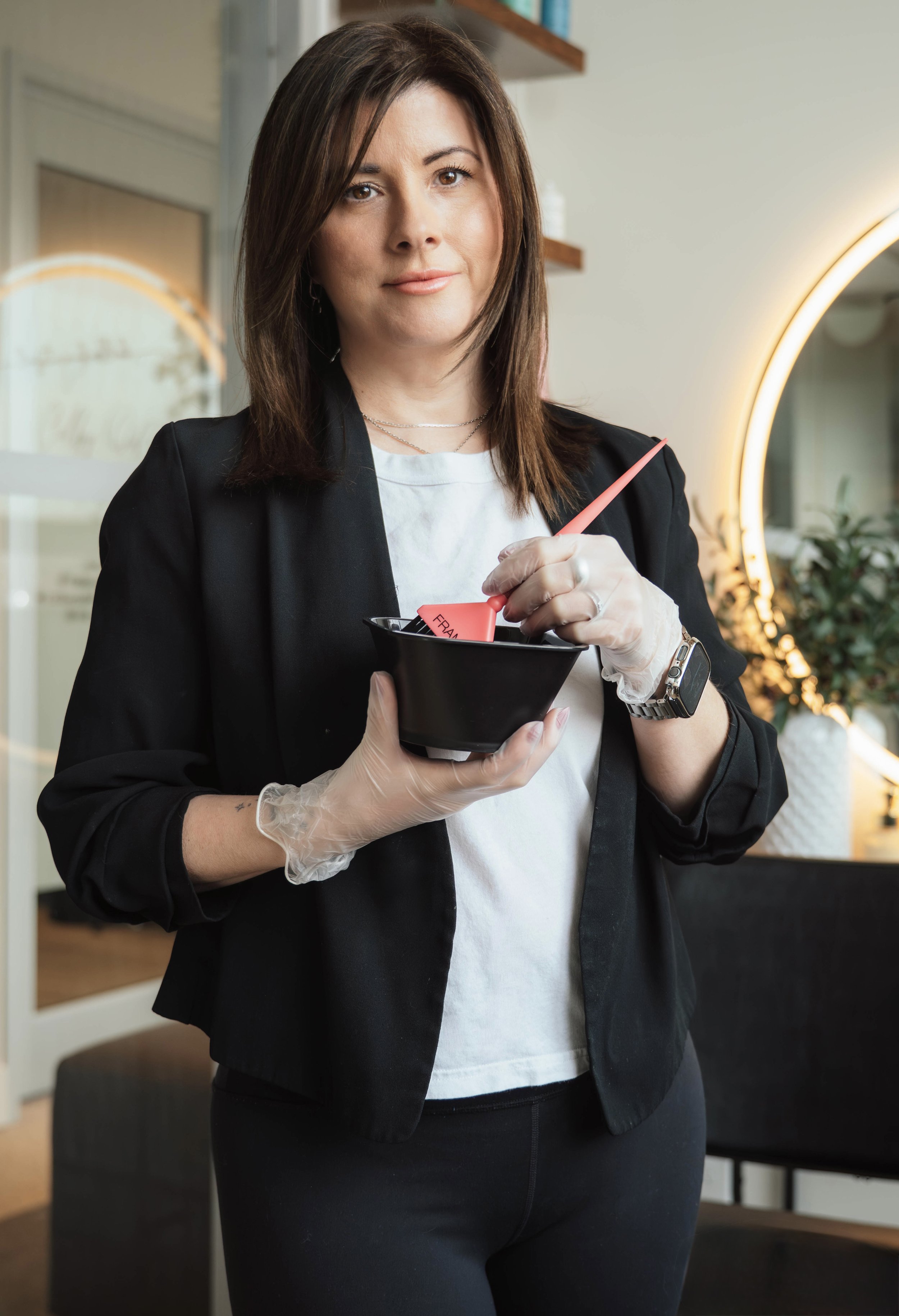 Woman holding a black bowl with pink plastic spatula, wearing a black blazer, white shirt, gloves, and a watch, inside a modern indoor space.
