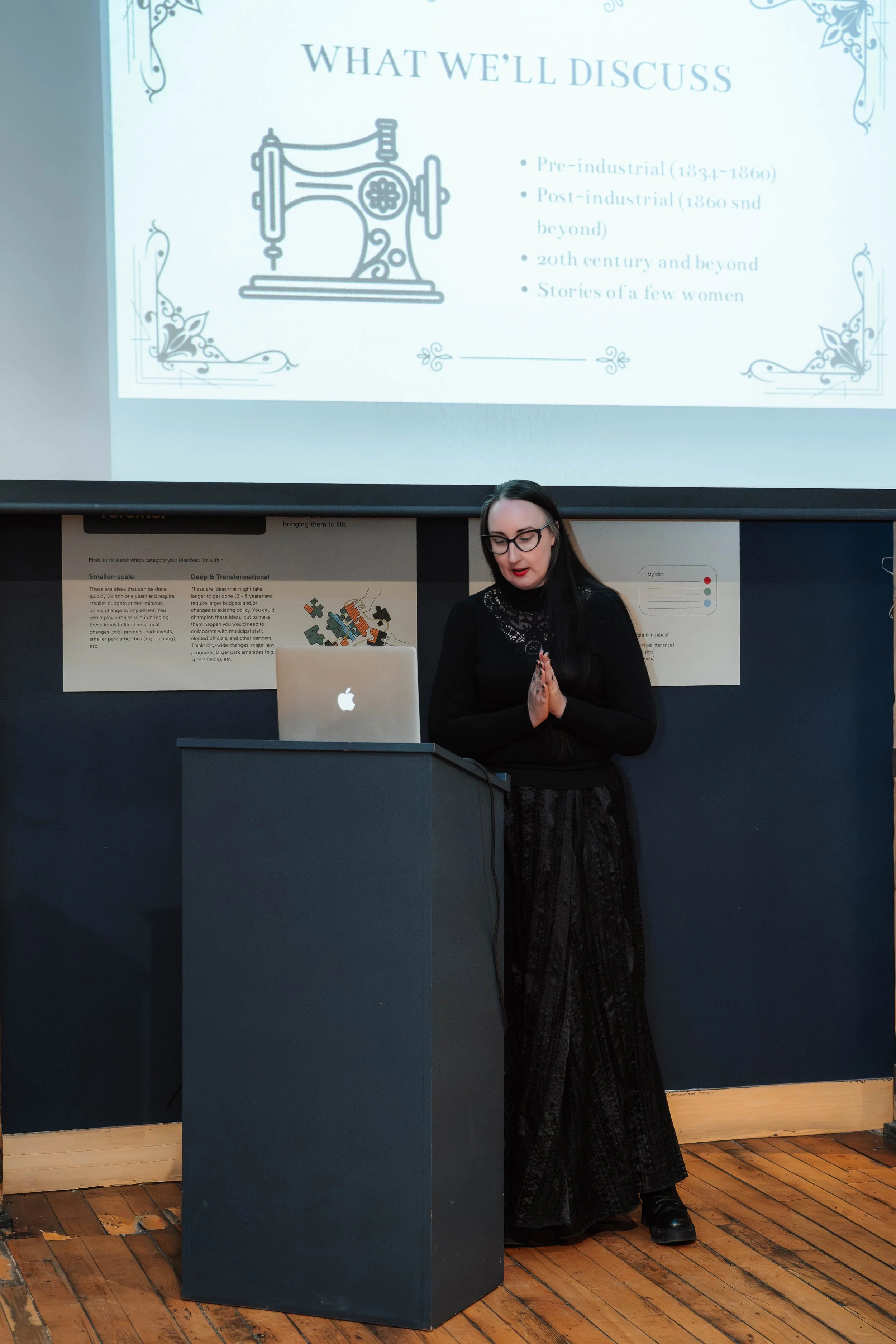 A woman with long dark hair and glasses giving a presentation in front of a large screen. The screen displays a slide titled 'What We'll Discuss' with a sewing machine illustration and bullet points about different historical periods, from pre-indust