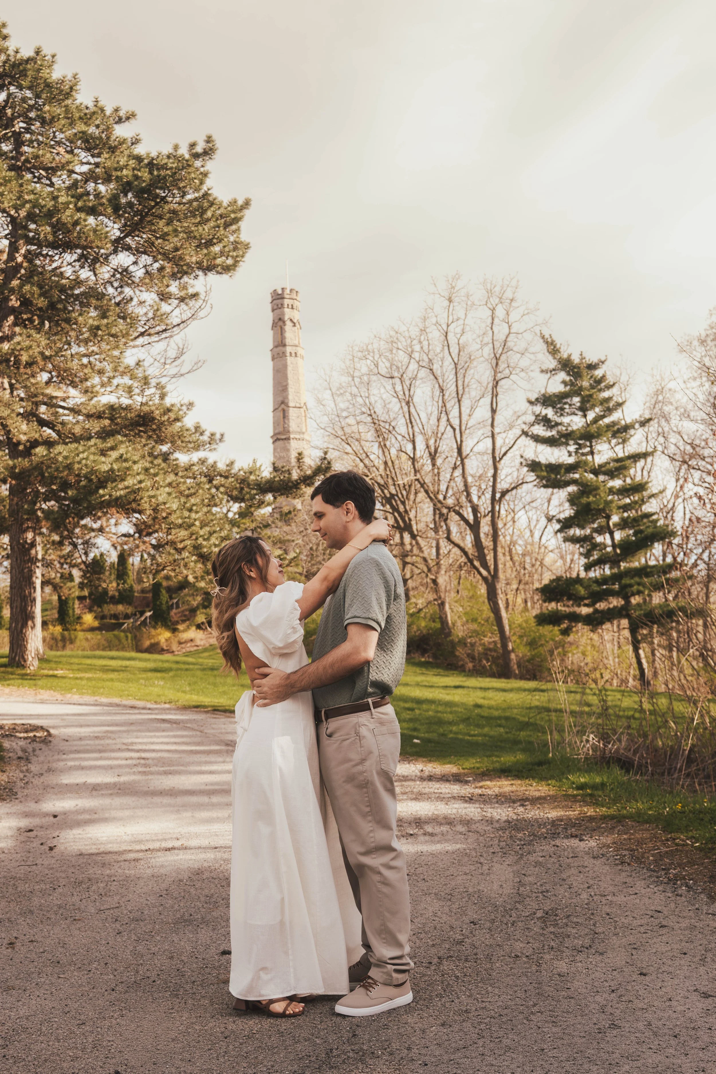 A couple dancing outdoors in a park with trees and a tall tower in the background during daylight.