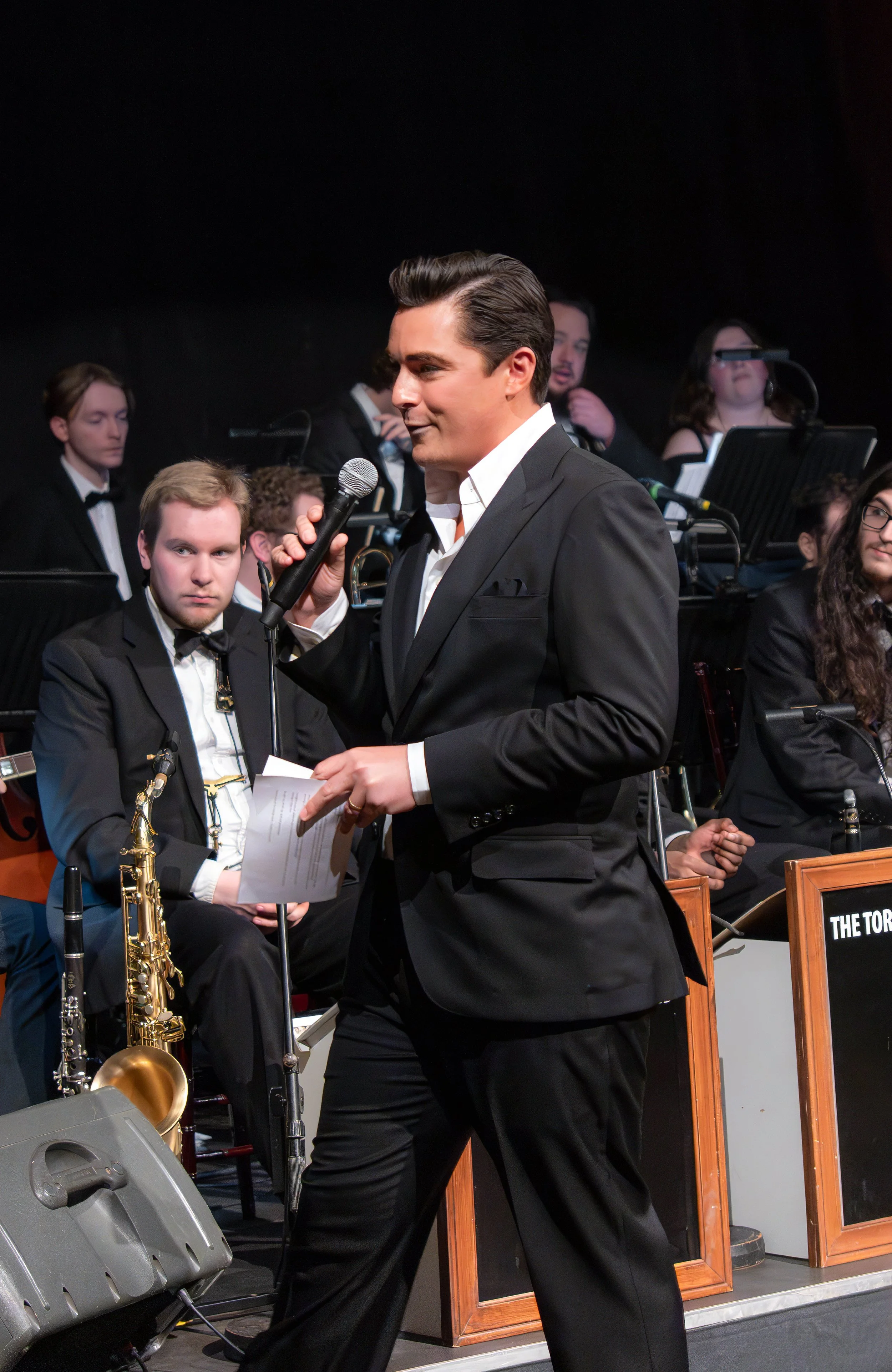 Man in black tuxedo speaking into a microphone at an orchestra concert.