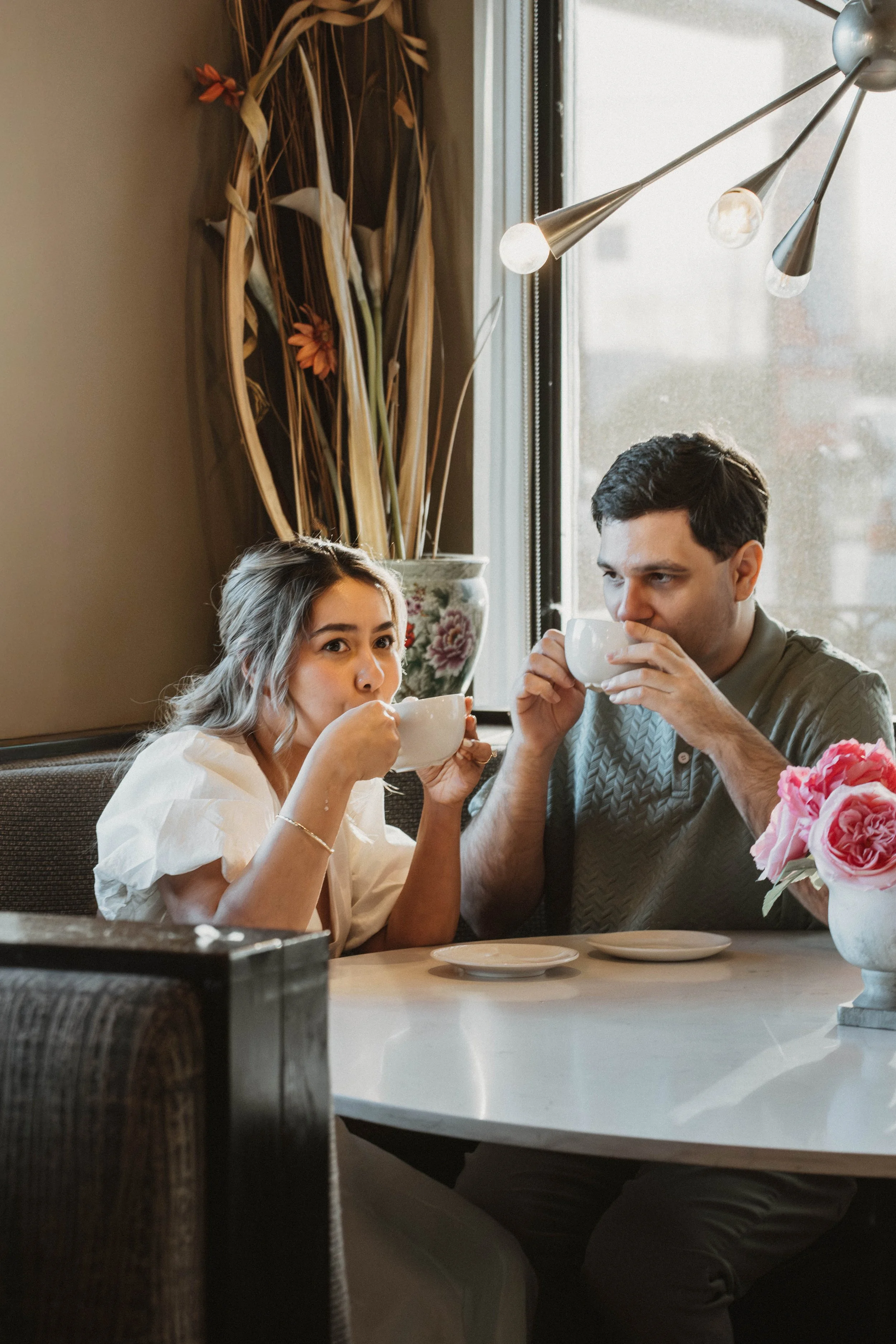 A young woman and a man are sitting at a table in a restaurant, drinking from white cups. The woman has blonde hair and is wearing a white blouse, while the man has dark hair and is wearing a gray shirt. There is a vase with pink roses on the table, 