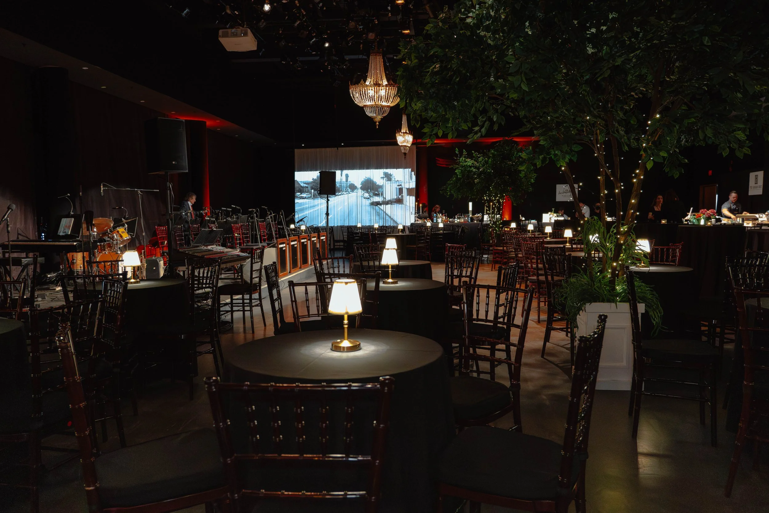 Interior of an elegant event venue with round tables covered in black cloths, illuminated by small lamps, surrounded by dark wooden chairs. A stage with musical instruments and microphones is visible on the left, and a large window at the back shows 