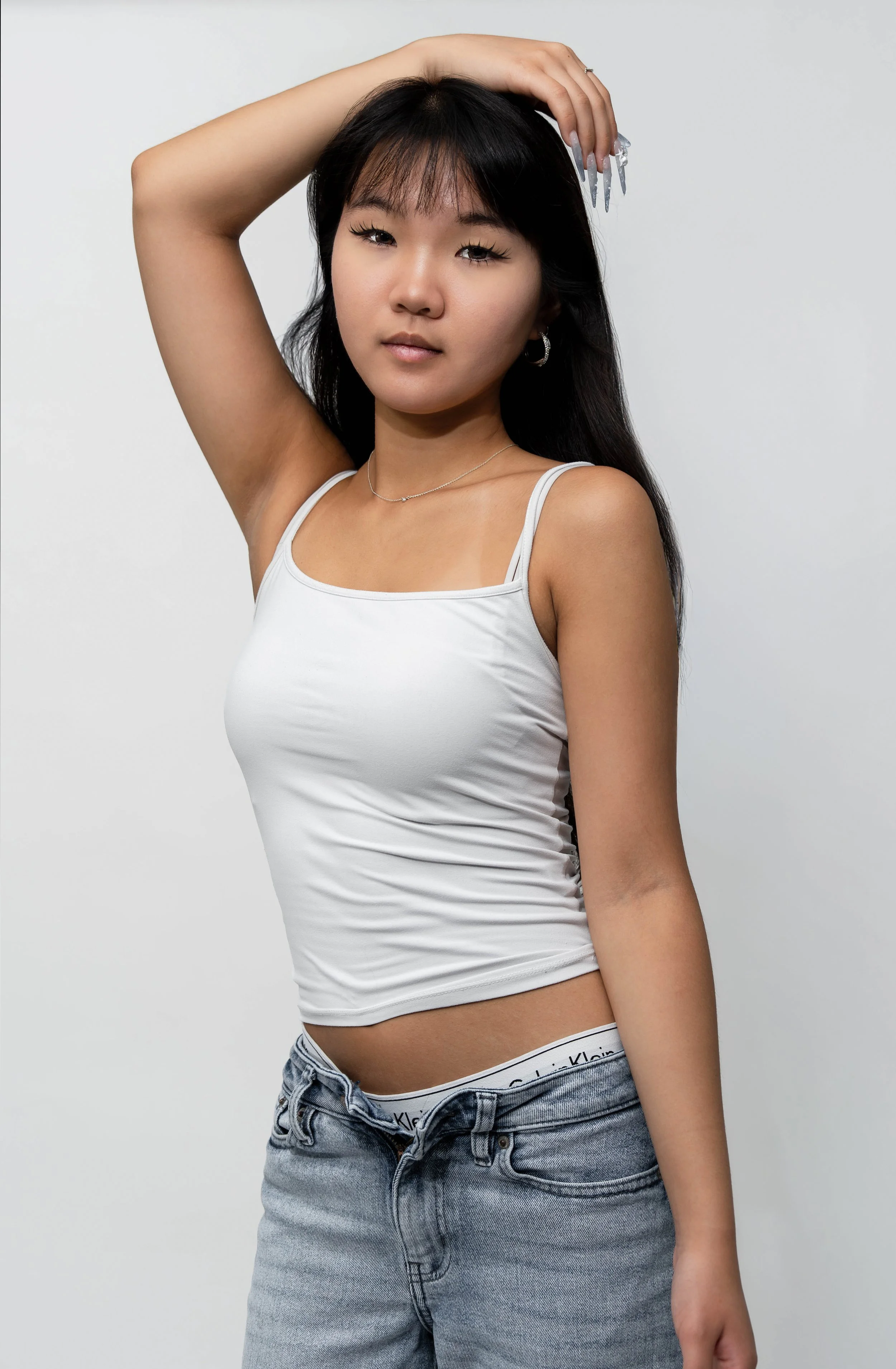 A young woman with long black hair, wearing a white tank top and blue jeans, posing with her left arm raised and hand resting on her head against a plain white background.