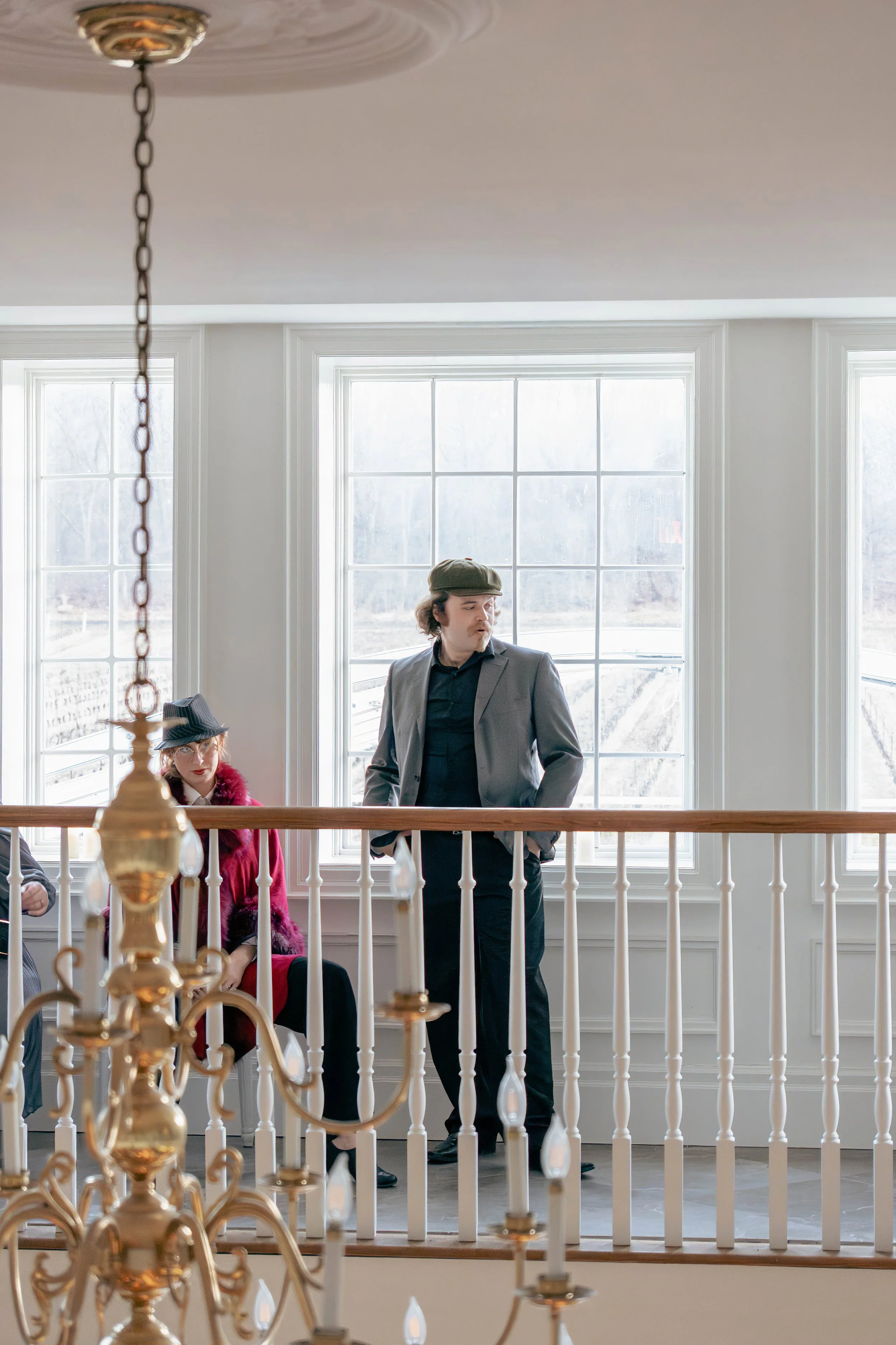 A man and a woman dressed in vintage clothing stand in a bright room with large windows. The man is standing and looking to the side, while the woman is seated next to him, wearing a hat and a red outfit. A chandelier is seen in the foreground, hangi