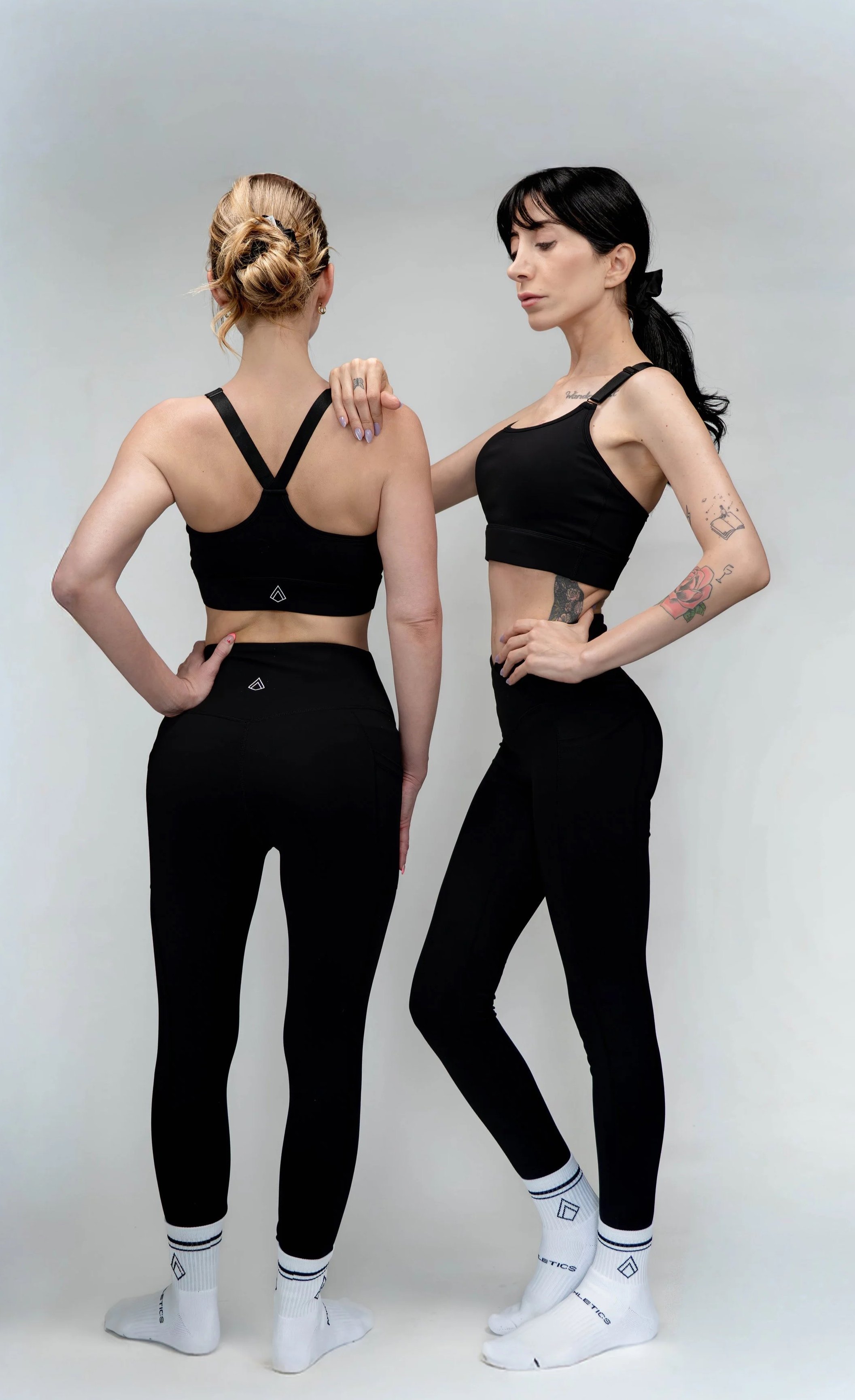 Two women in black athletic wear standing facing each other with one woman touching the other's shoulder. They are wearing matching black tops, black leggings, and white socks, against a plain white background.