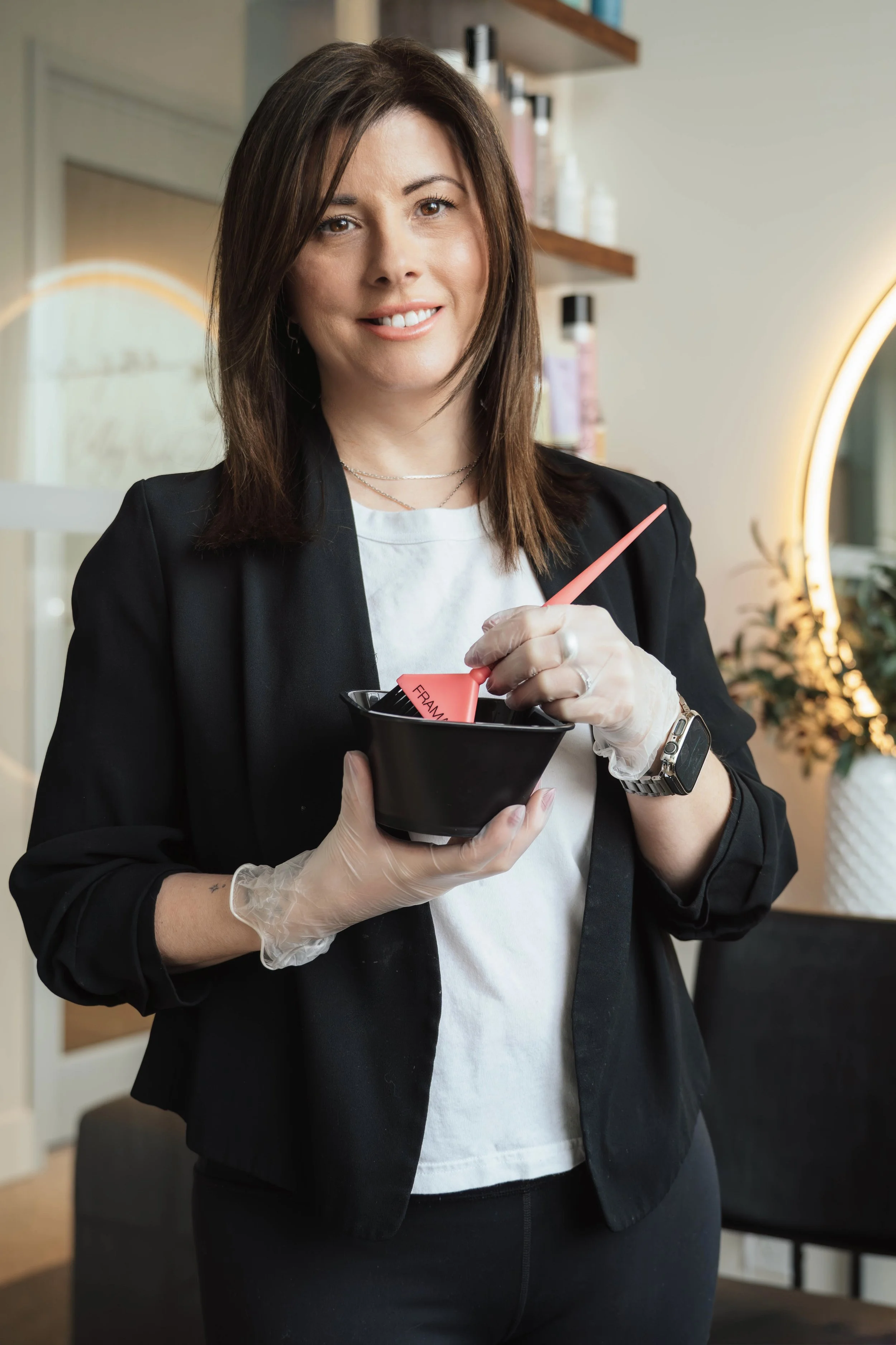 A woman with shoulder-length brown hair, wearing a black blazer, white T-shirt, and a smartwatch, is holding a black bowl and using a pink spatula. She is indoors, possibly in a salon or spa, with a wall-mounted shelf and a round mirror glowing in th