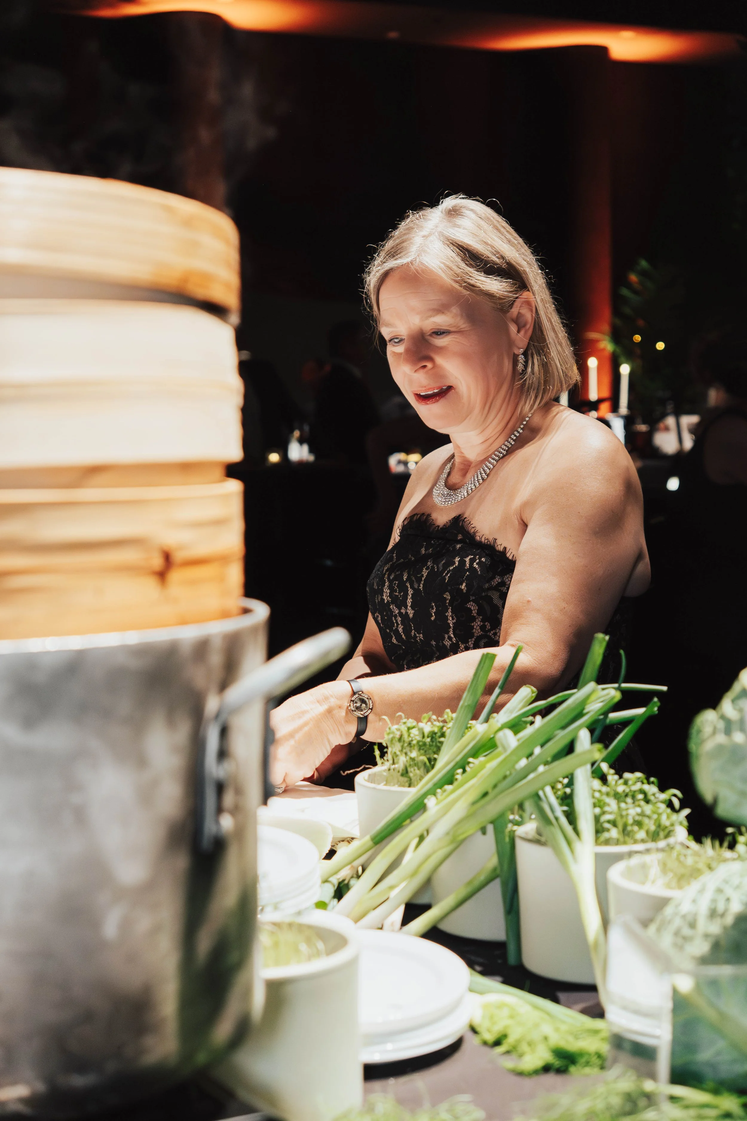 An older woman with blonde hair dressed in a black lace strapless dress and jewelry, standing at a table with cups, plates, and fresh green vegetables in white cups, in a dimly lit setting.
