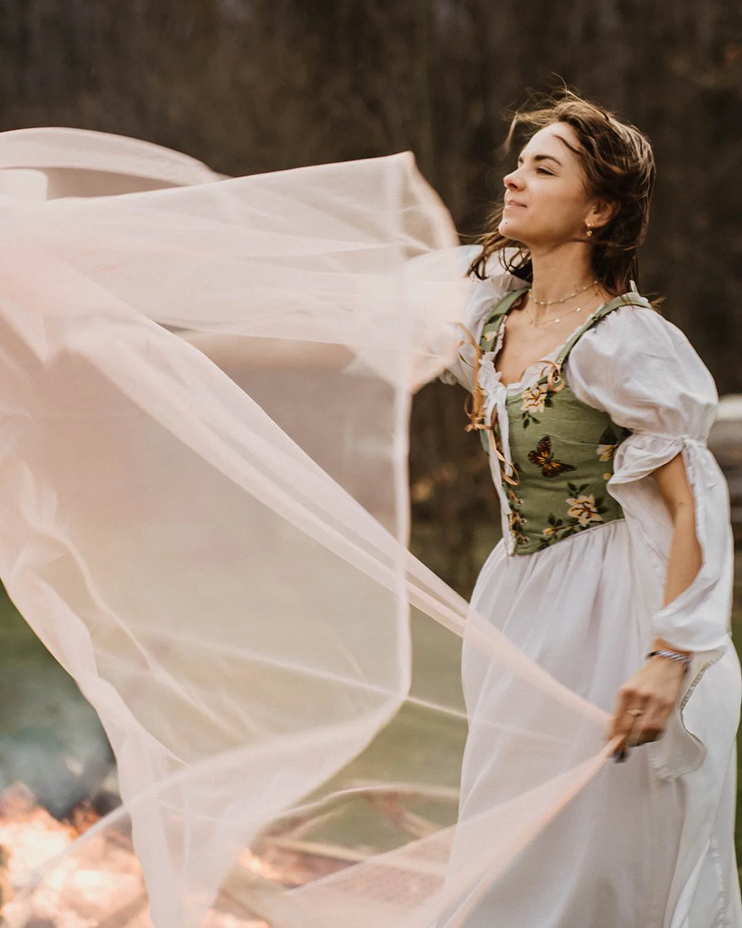 A young woman with short brown hair wearing a vintage dress with puffy sleeves and floral embroidery in a natural outdoor setting, holding a flowing light-colored fabric.