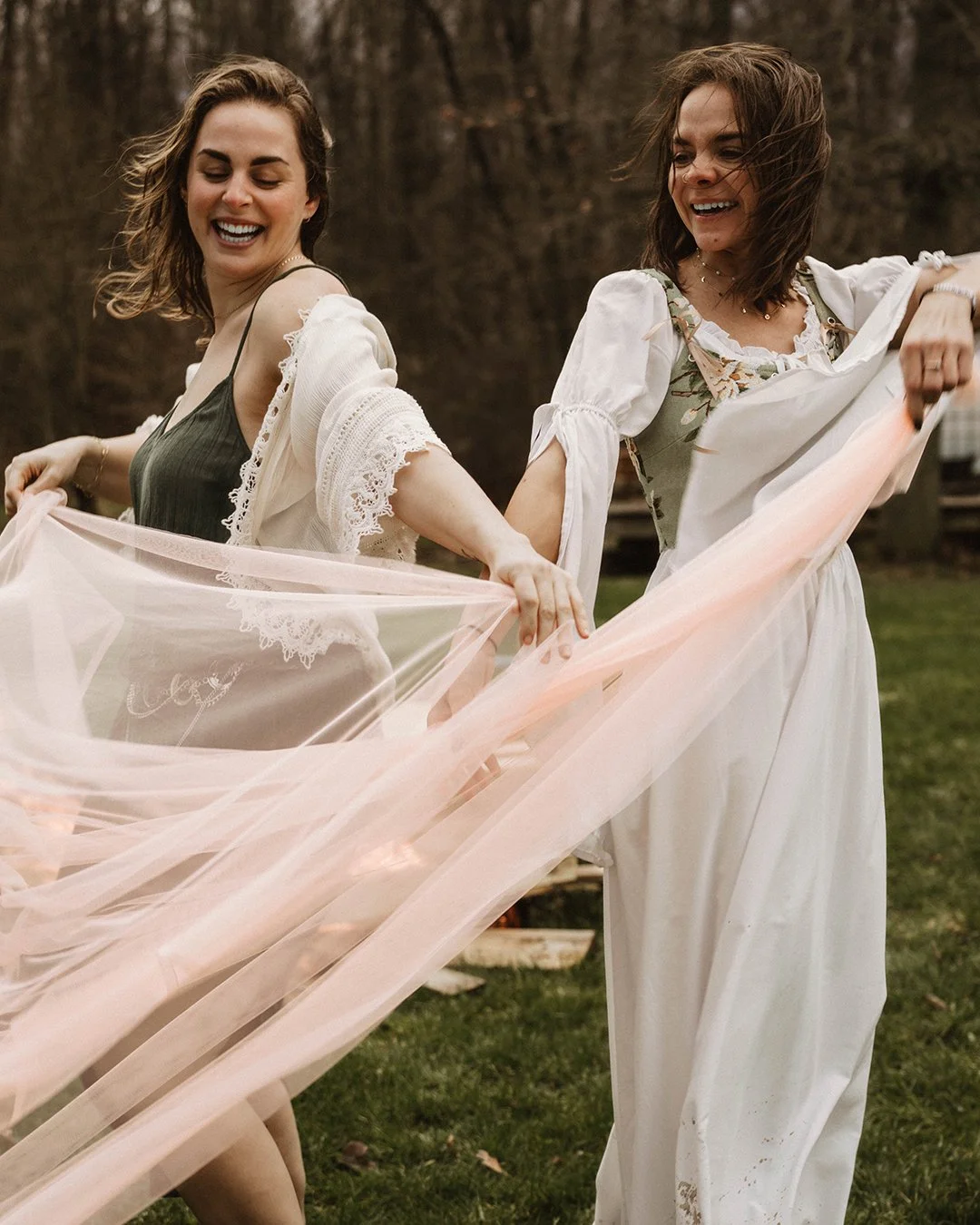 Two women dancing outdoors on grass, smiling and holding a flowing piece of pink fabric between them. The background is a wooded area.