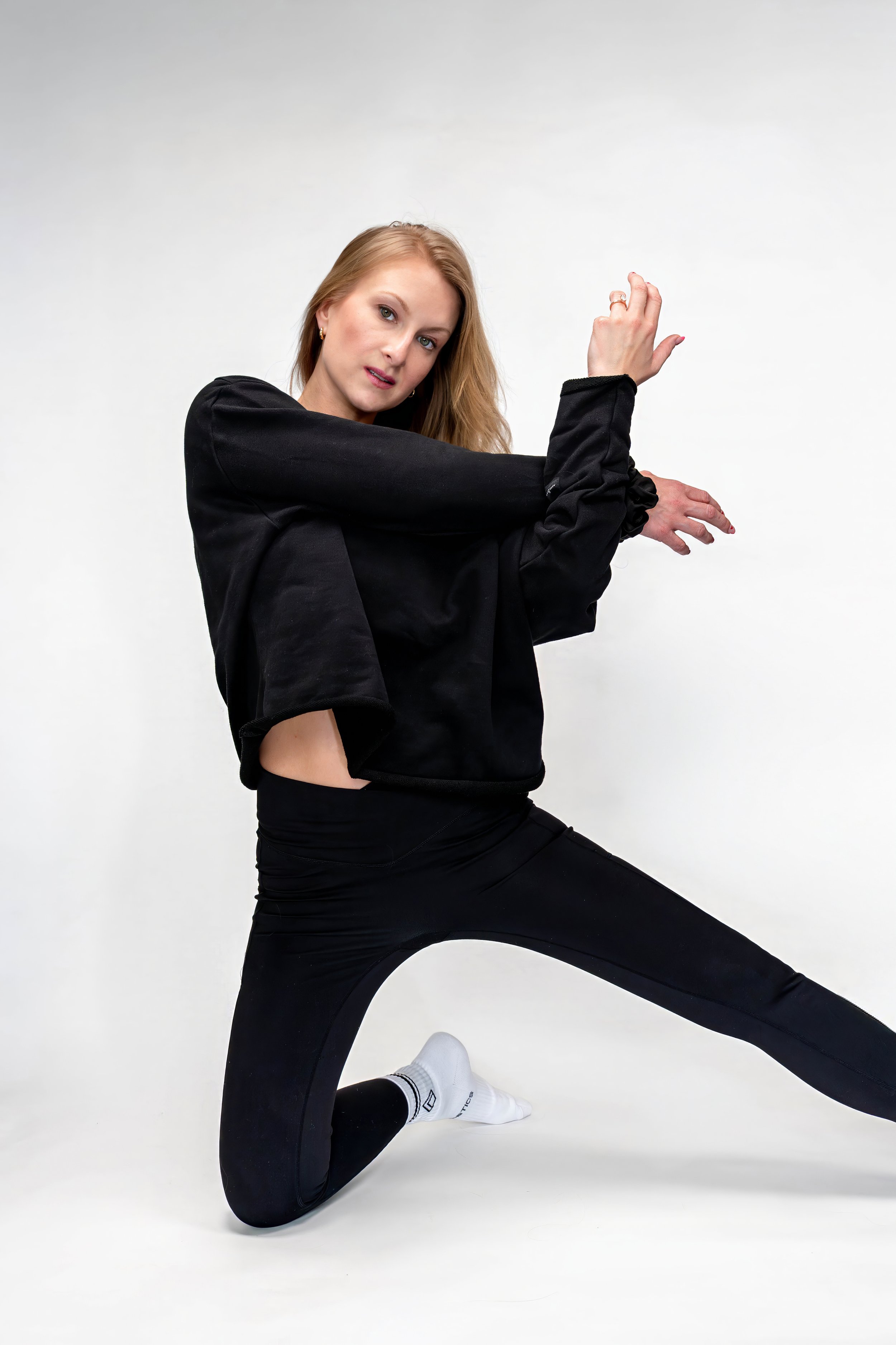 A woman with long blonde hair practicing dance or fitness moves on a white background, wearing black workout clothes and white socks.
