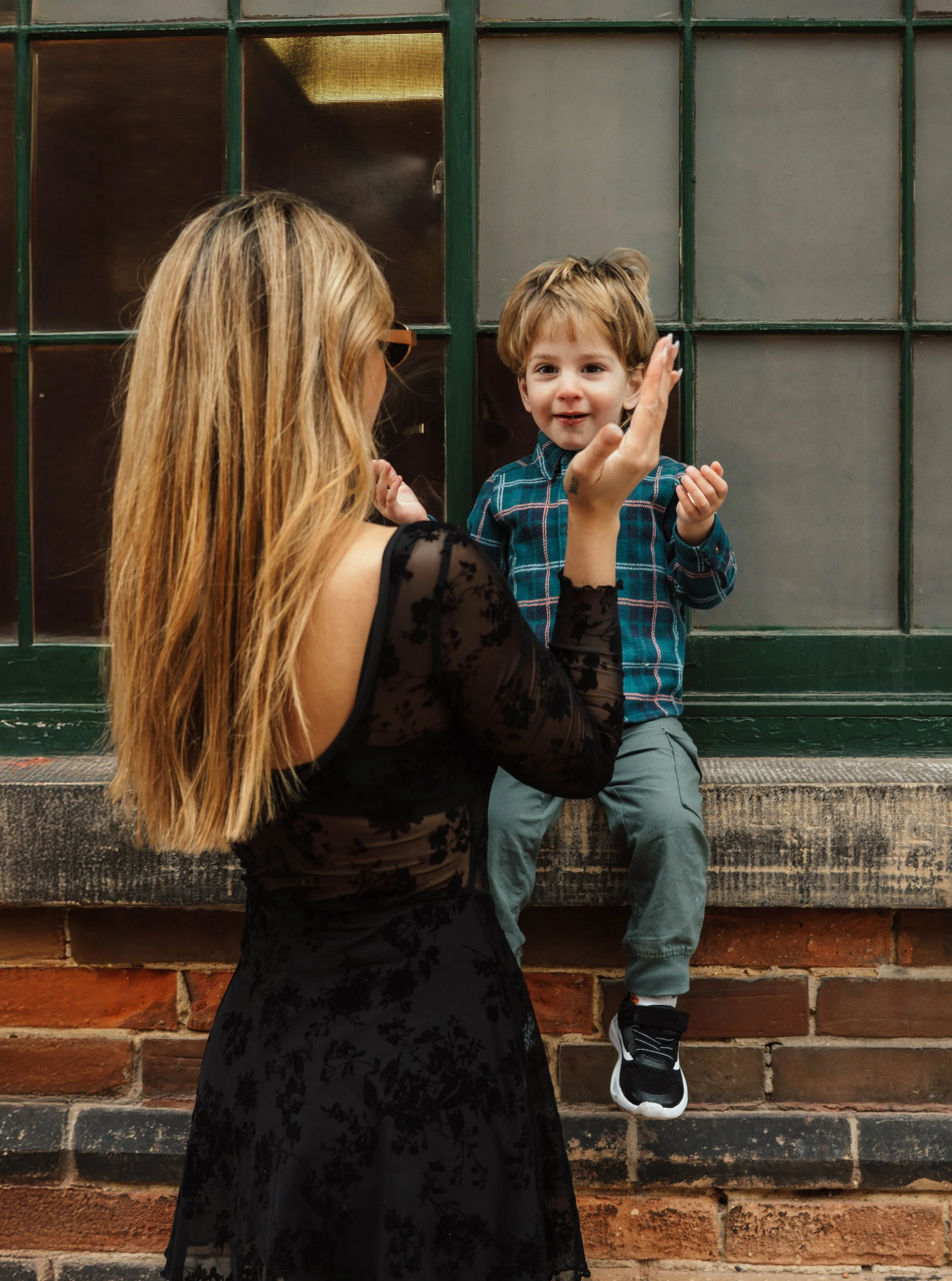 A woman with long blonde hair and wearing a black lace dress is interacting with a young boy. The boy, with light brown hair, is sitting on a brick ledge against a green-framed glass window. He is wearing a blue plaid shirt, gray pants, and black sne