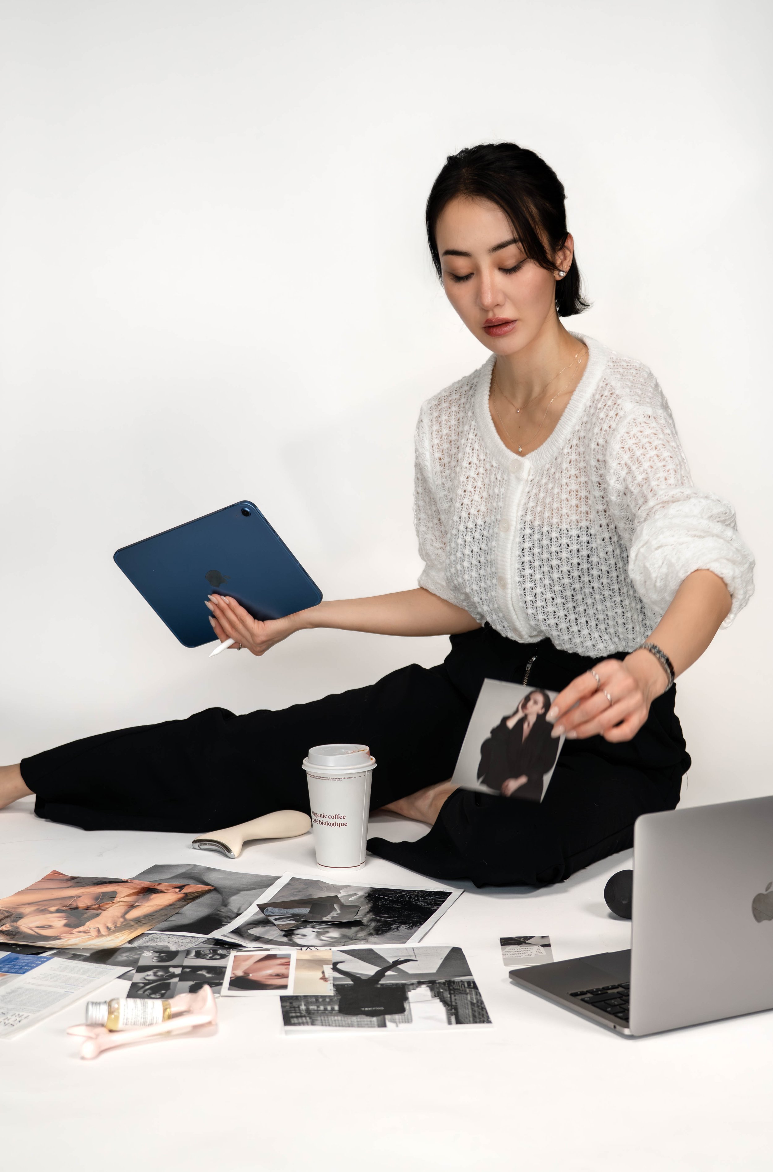 A woman sitting cross-legged on the floor, holding a photograph and a tablet, surrounded by photographs, a laptop, a coffee cup, and personal items in front of a white background.