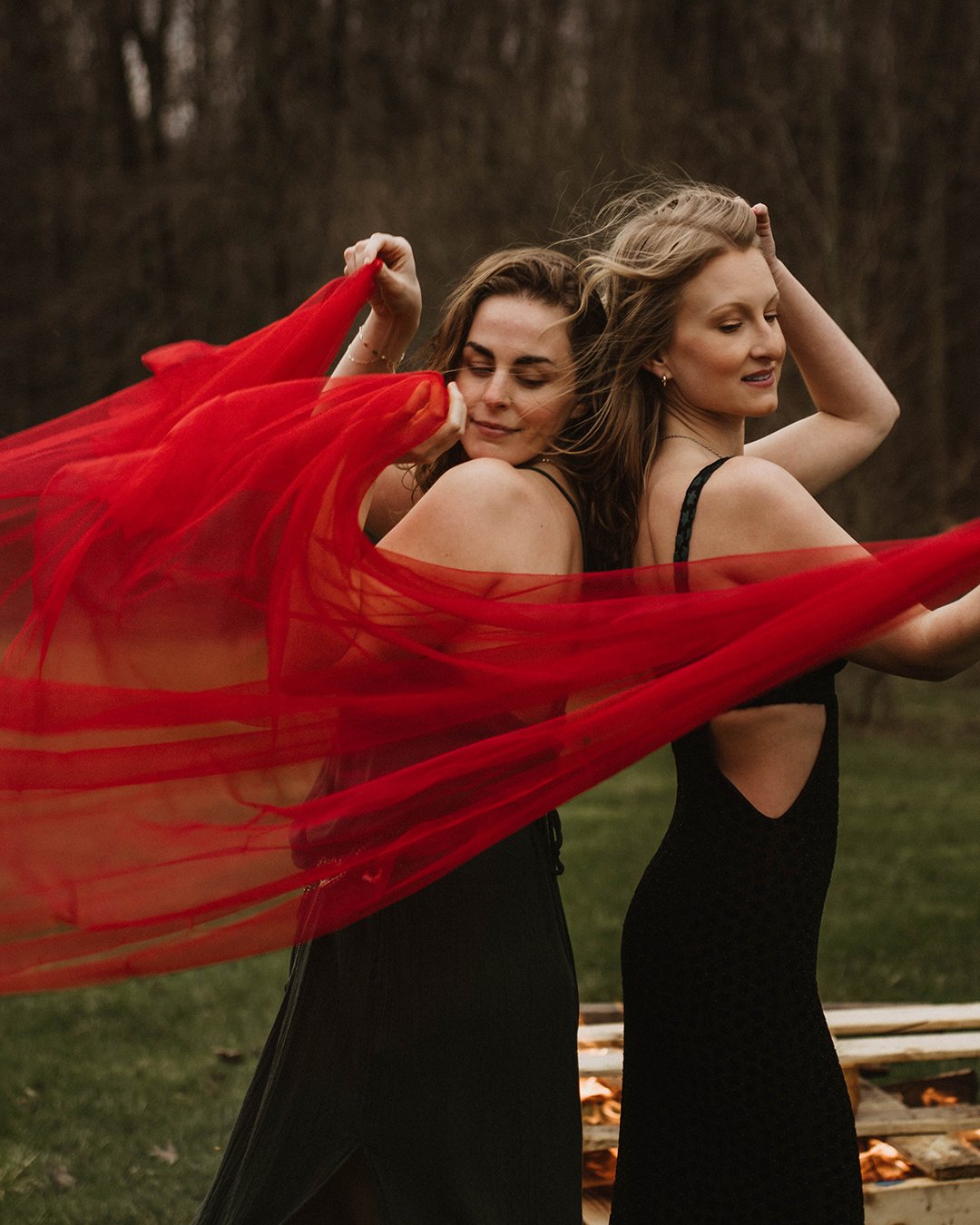 Two women in black dresses, one holding a flowing red sheer fabric, standing outdoors with a fire burning in the background.