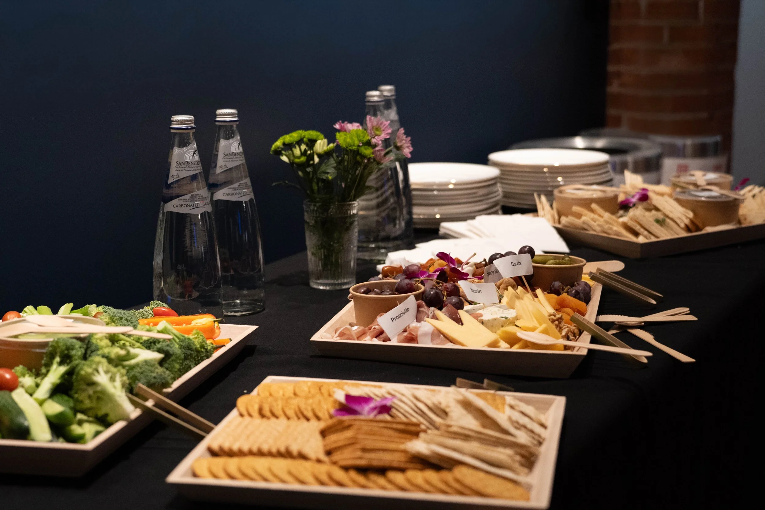 A table set with an assortment of food and drinks, including trays of vegetables, cheeses, grapes, crackers, and spreads, with bottles of sparkling water, stacked plates, napkins, and flowers in the background.