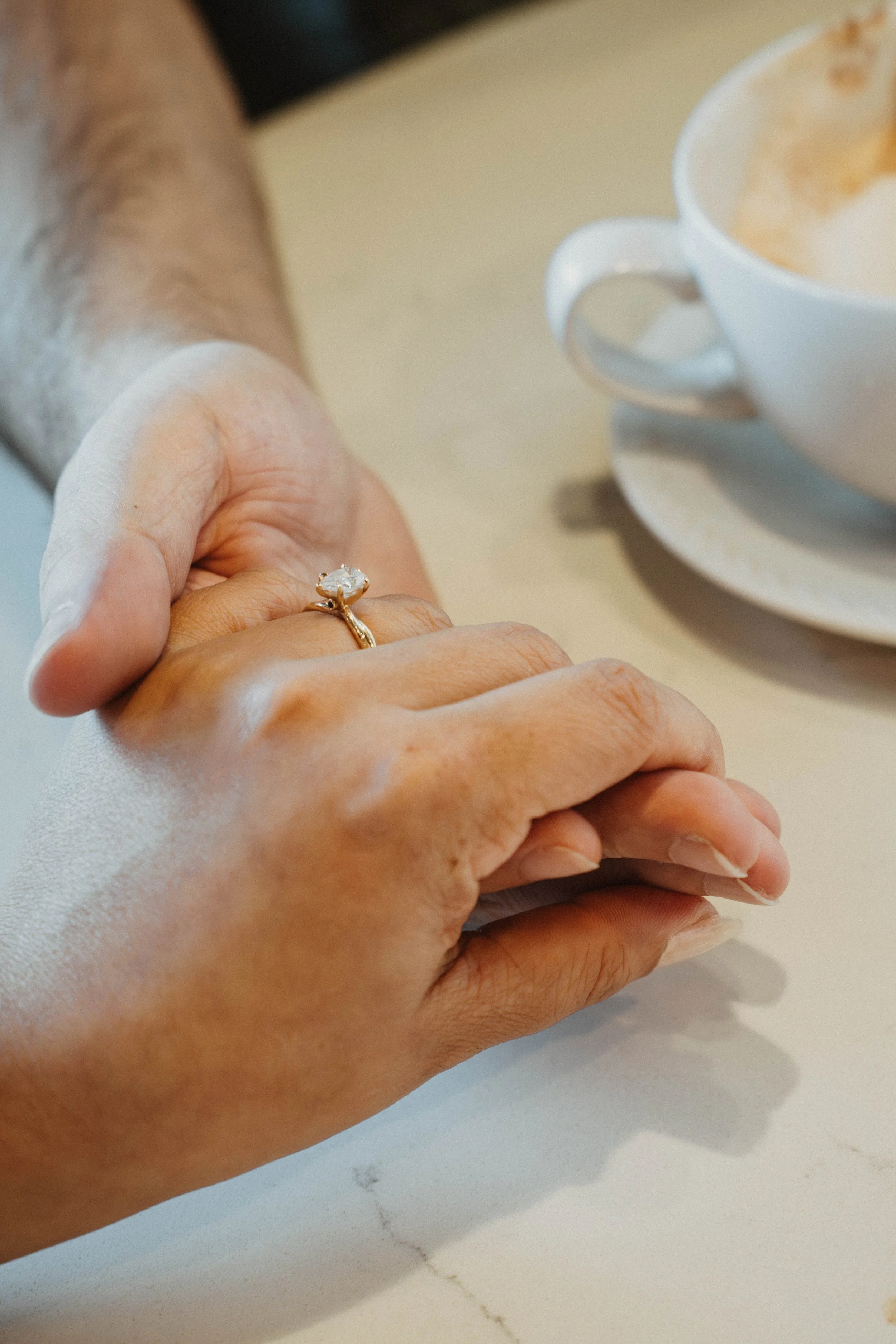 Close-up of two hands holding each other, with an engagement ring featuring a large gemstone on the ring finger. In the background, there is a white ceramic cup and saucer with a beverage, possibly coffee or tea, on a light-colored surface.