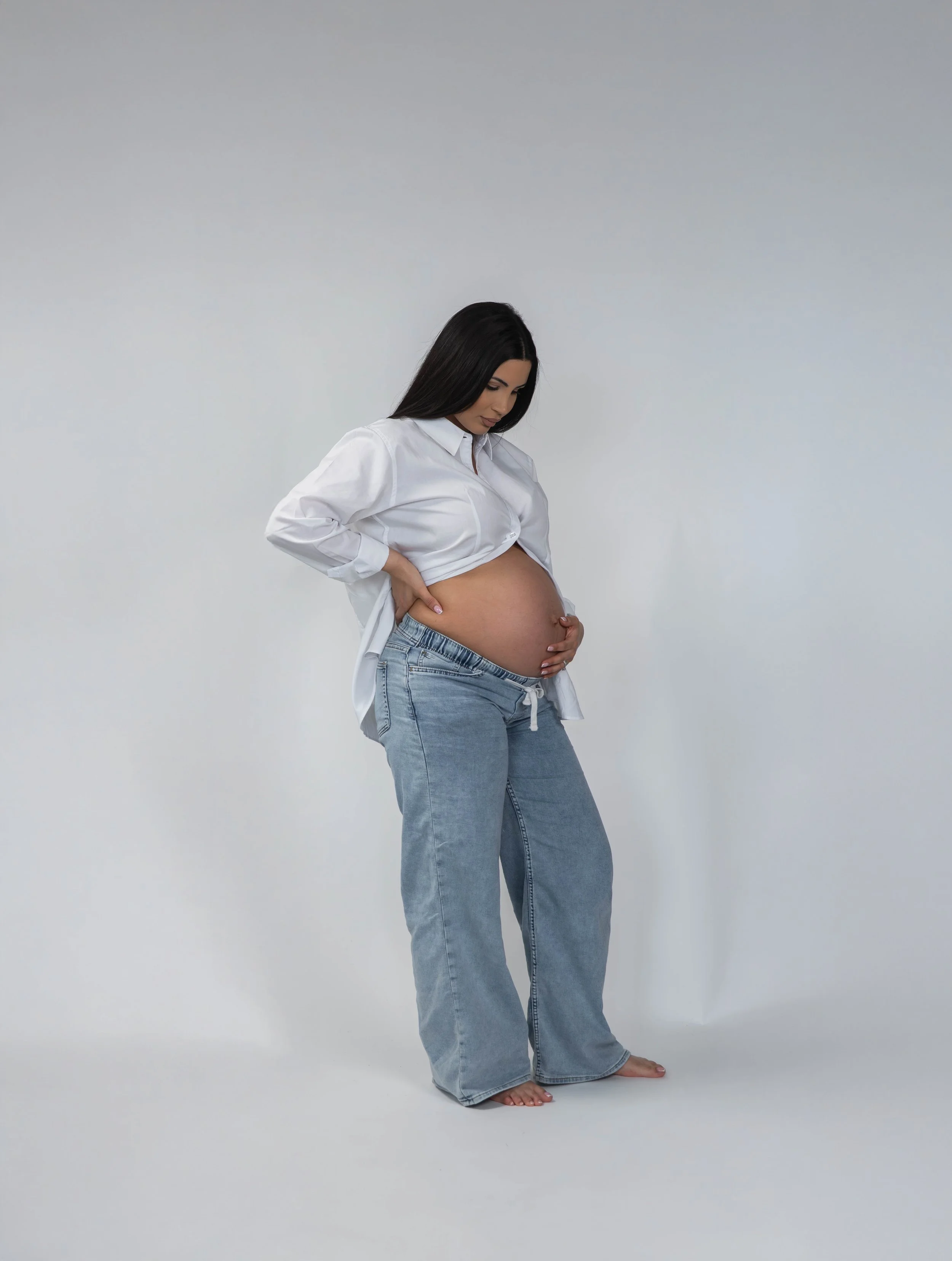 A pregnant woman with long black hair, wearing a white shirt and jeans, standing barefoot against a plain wall, looking down at her belly and gently holding it with both hands.