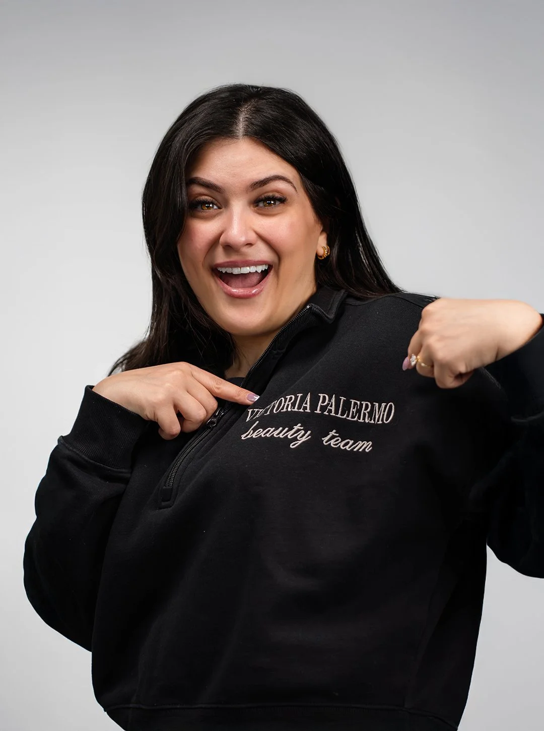 A smiling woman with dark hair, wearing a black zip-up sweatshirt that says 'Victoria Palermo beauty team,' pointing at the logo and flexing her arm.