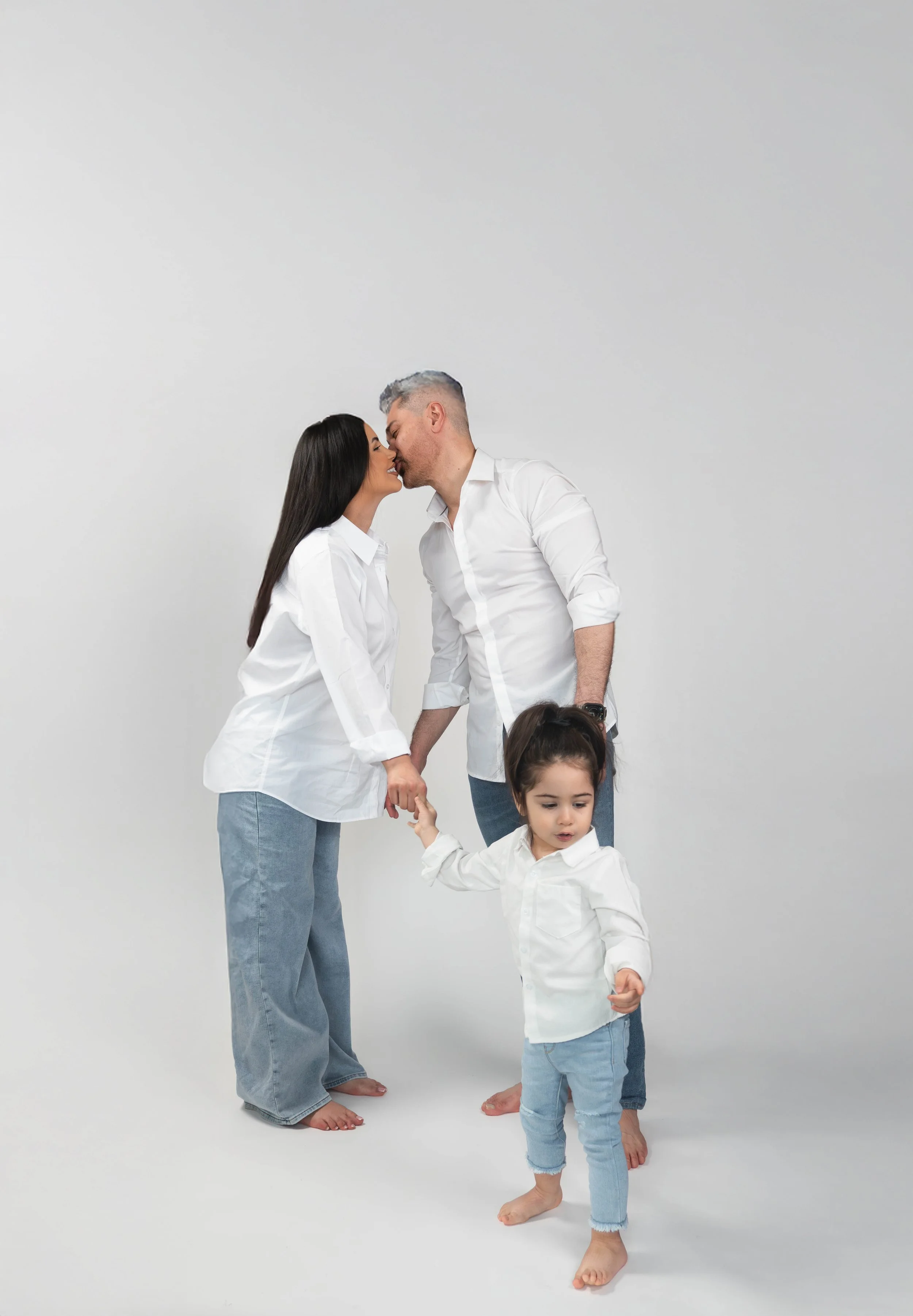 A family of four holding hands and sharing a kiss against a plain white background. The adults are barefoot, wearing white shirts and blue jeans, while the child in the foreground is also barefoot, dressed in a white shirt and light blue jeans.