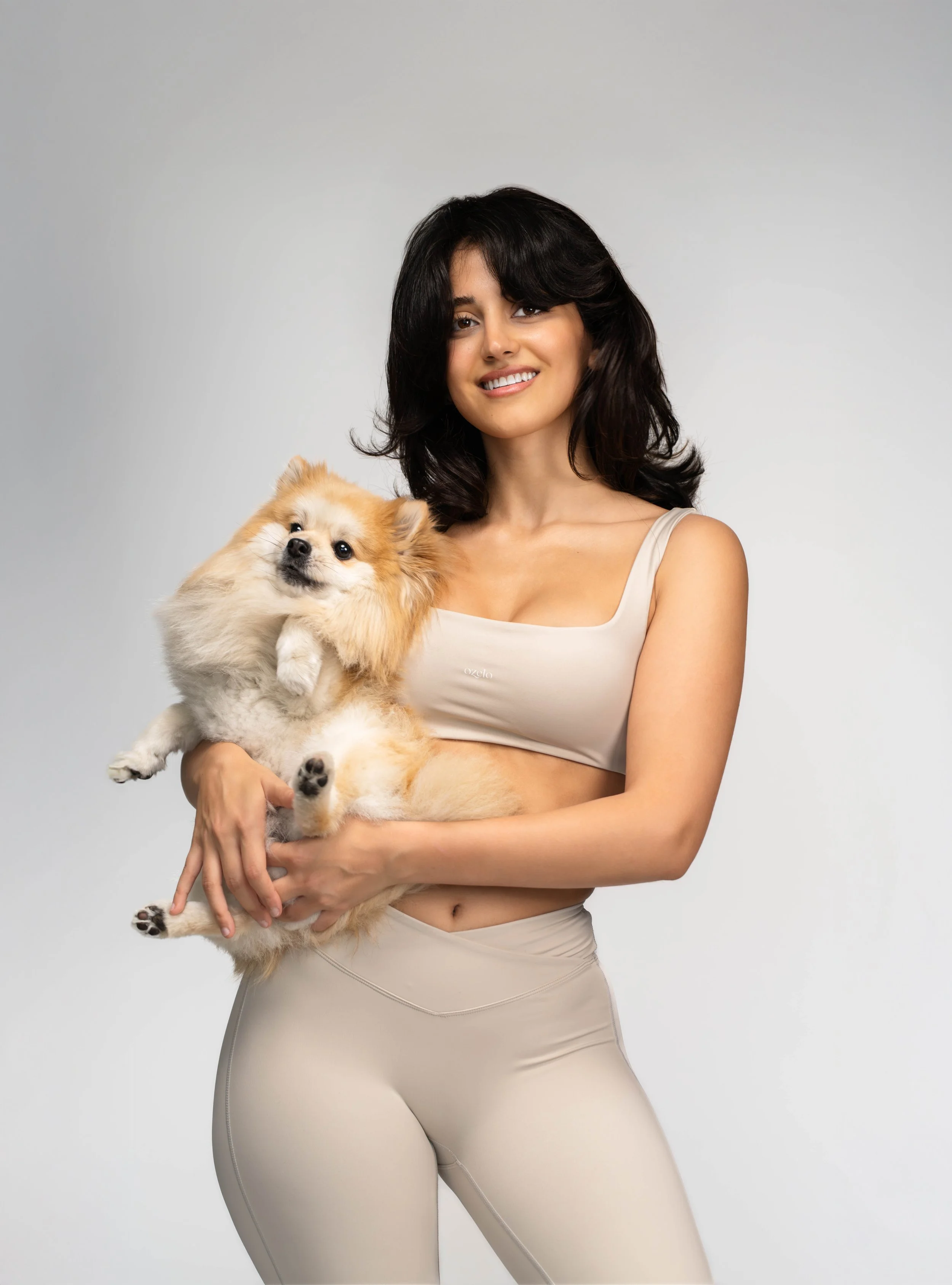 A woman with dark hair smiling and holding a small, fluffy dog against a plain light gray background.