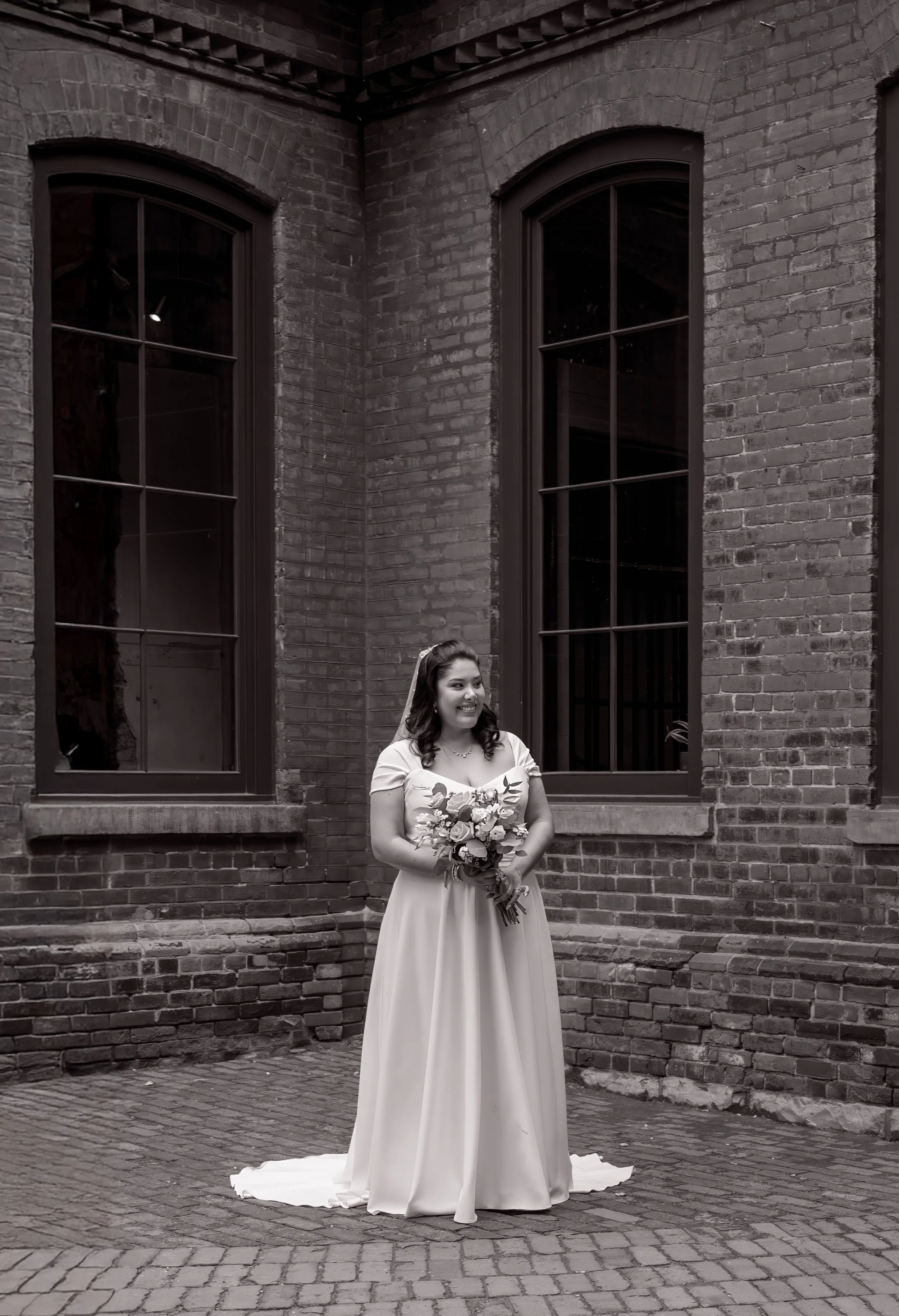 A woman in a wedding dress holding a bouquet, standing in front of a brick building with two large windows, smiling.