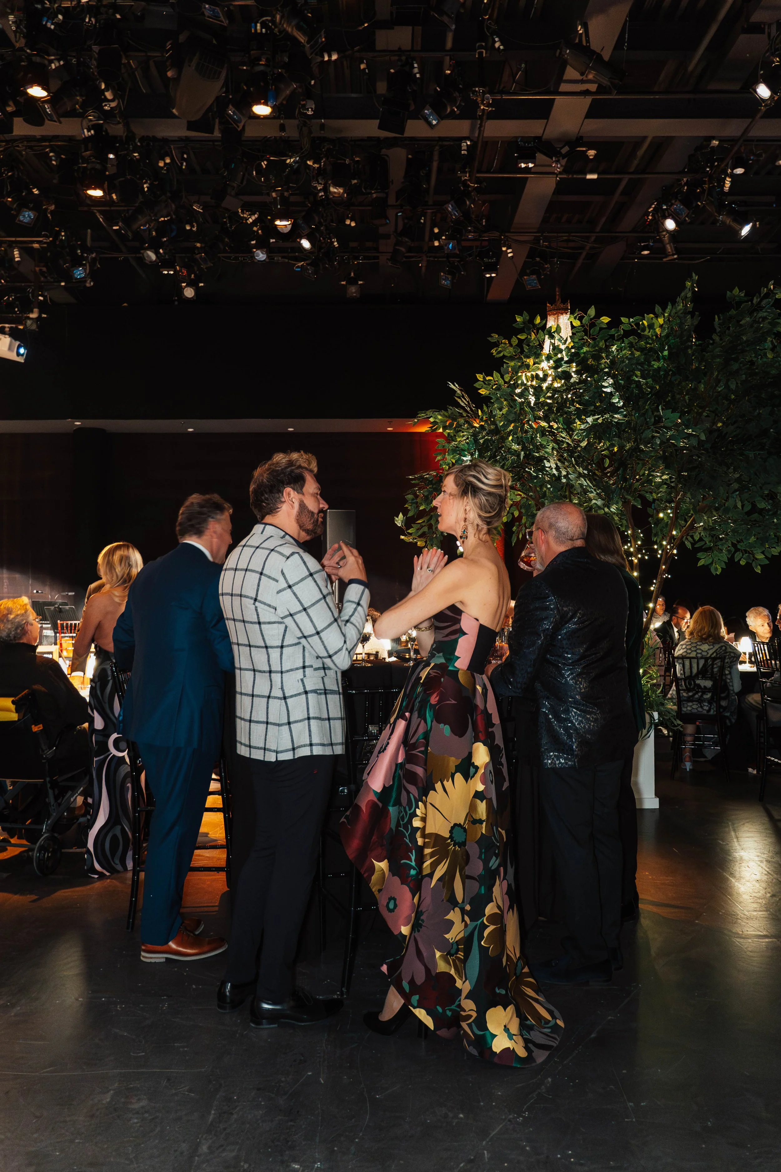 People talking at a formal event in a decorated venue with dim lighting, a large tree with string lights, and seated guests in the background.