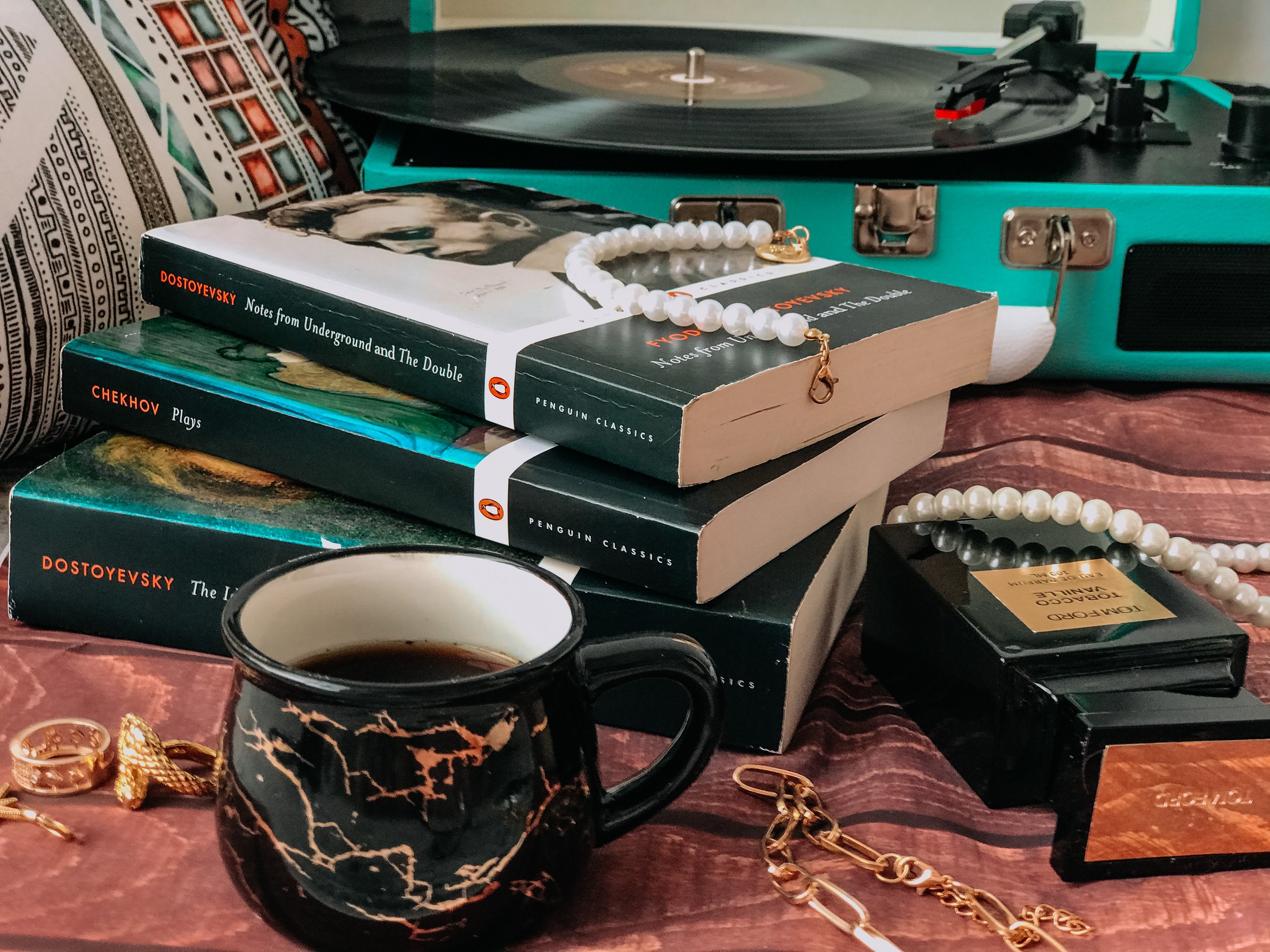 An assortment of books, a mug with a dark liquid, jewelry including pearl necklaces and gold chains, a vintage record player, and a black box perfume on a wooden surface.