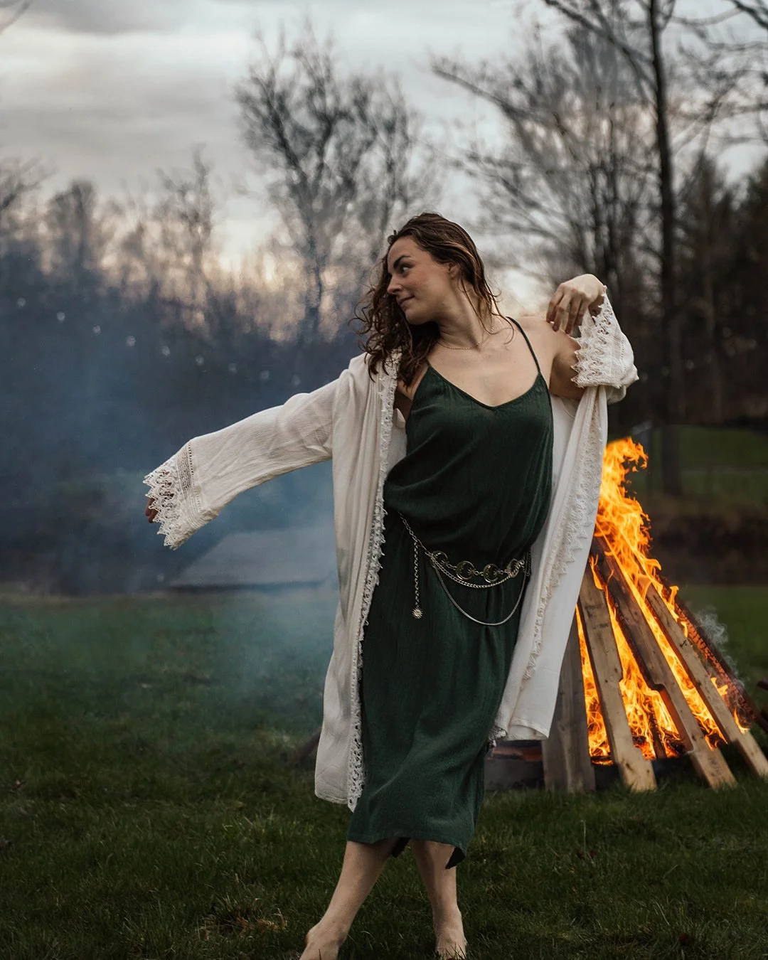 A woman in a green dress and white lace robe dances near a bonfire on a grassy field in a park, with bare trees and a cloudy sky in the background.