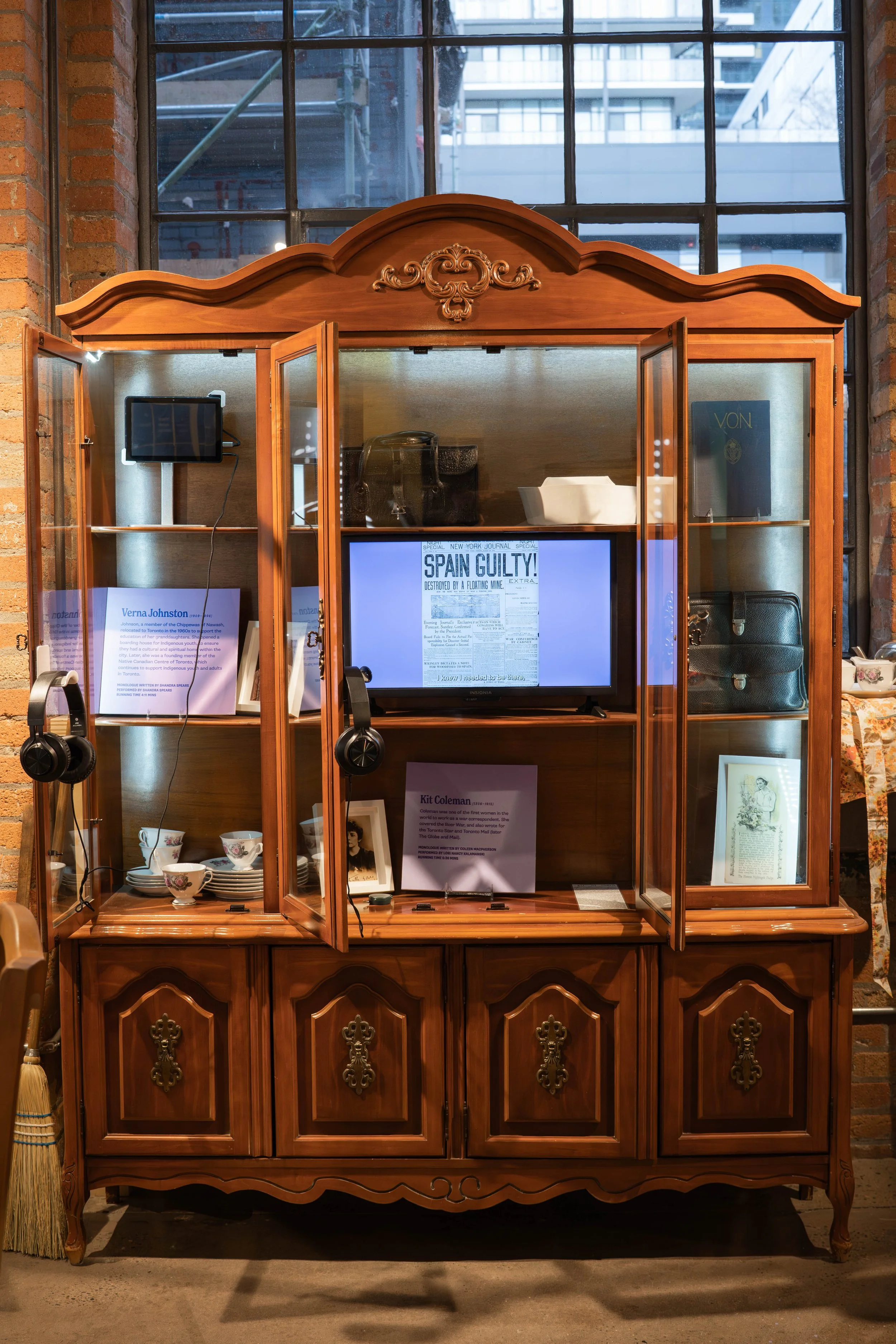 A wooden display cabinet containing various items including a small television, framed documents and photos, teacups, and a leather bag, with a large window in the background.