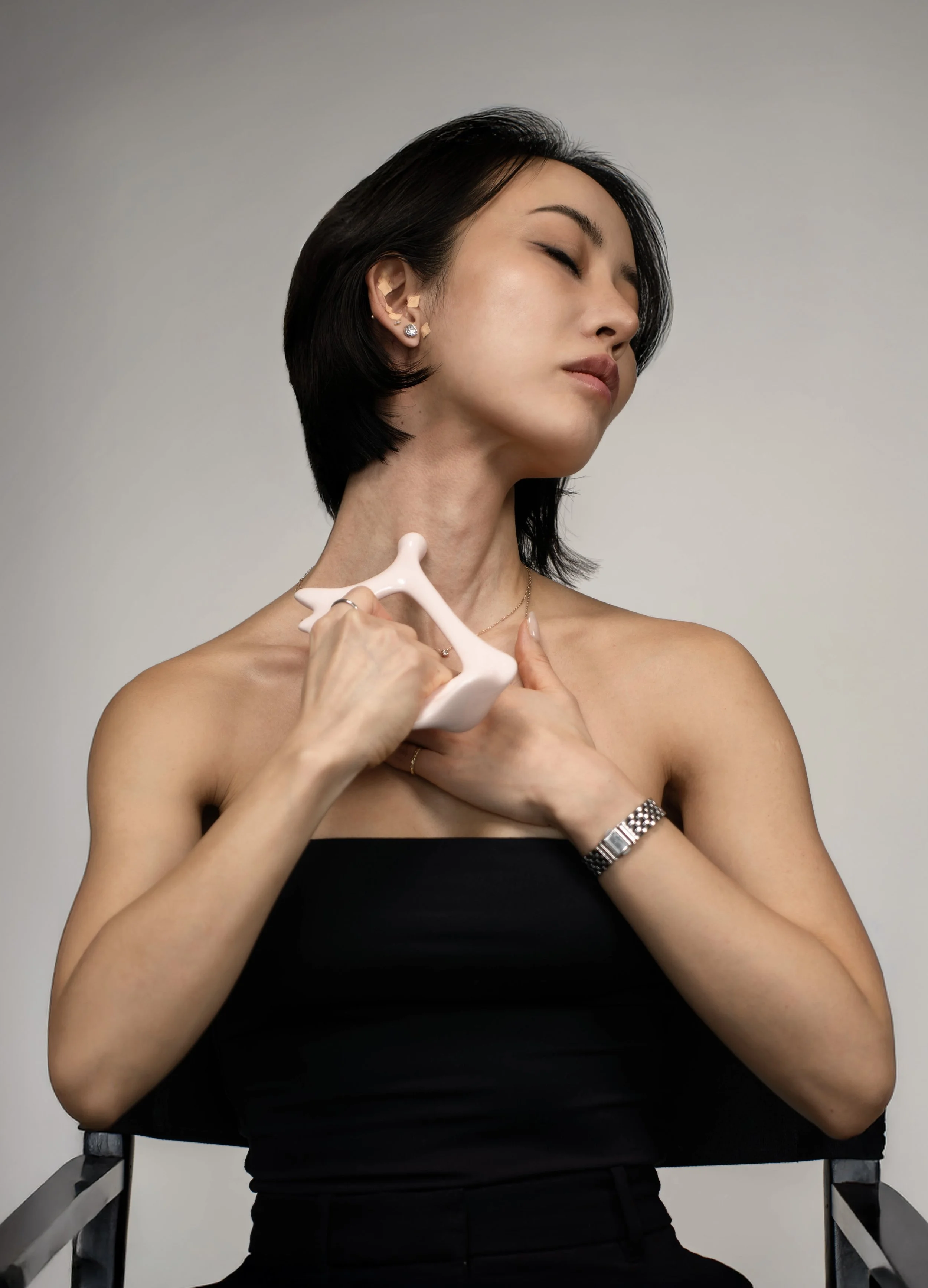 Woman with short black hair applying gua sha massage on her neck using a white stone, dressed in a strapless black top, against a plain background.