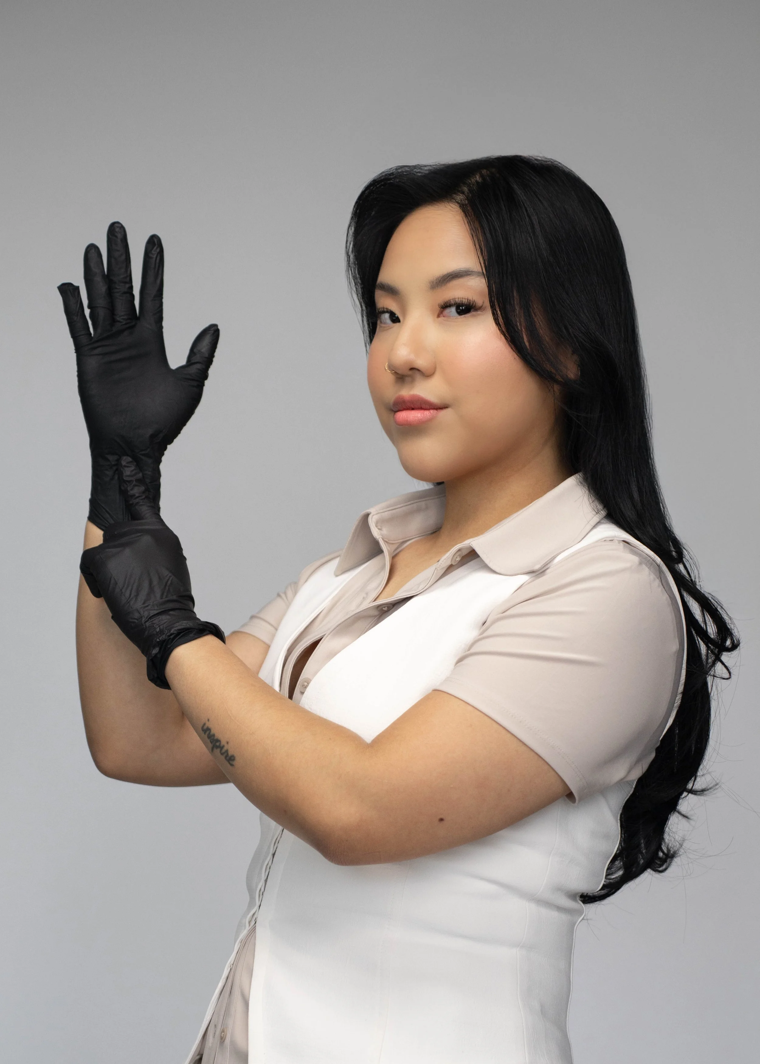 A woman with black hair wearing black gloves and a beige and white outfit, standing against a gray background, looking at the camera, with her right hand raised as if waving.