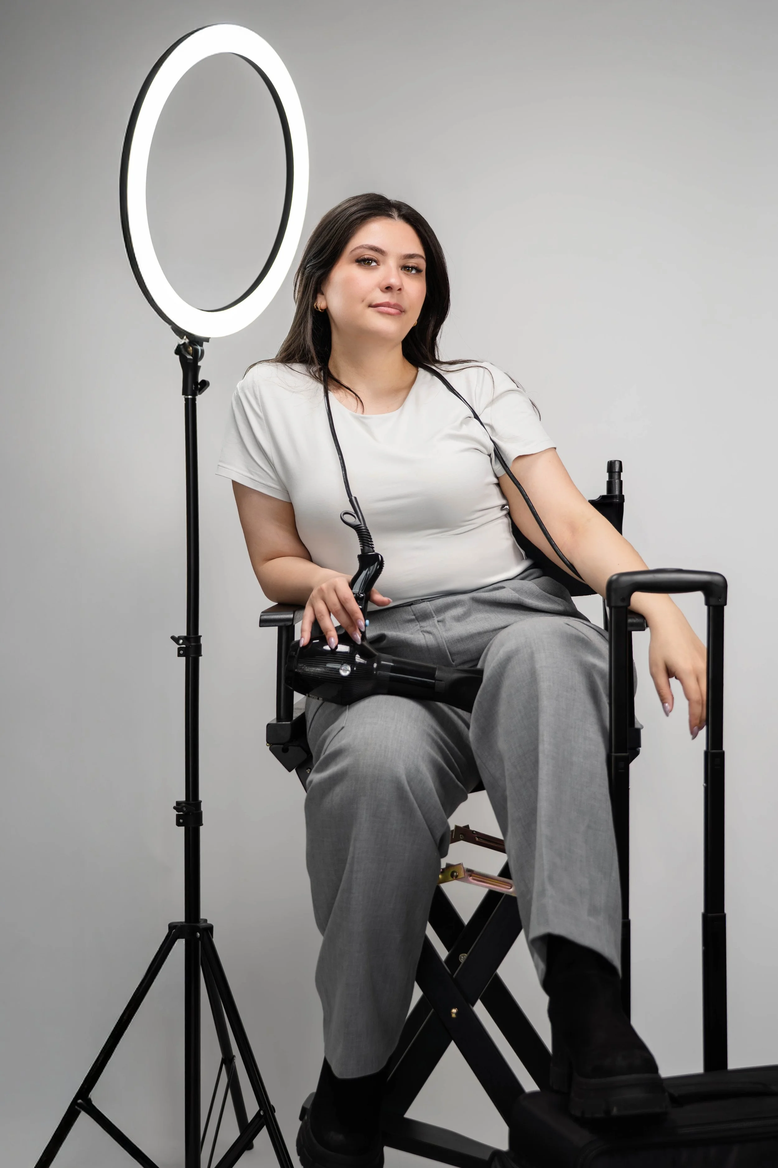 Woman sitting in a motorized wheelchair with a ring light spotlighting her, wearing a white top and gray pants.