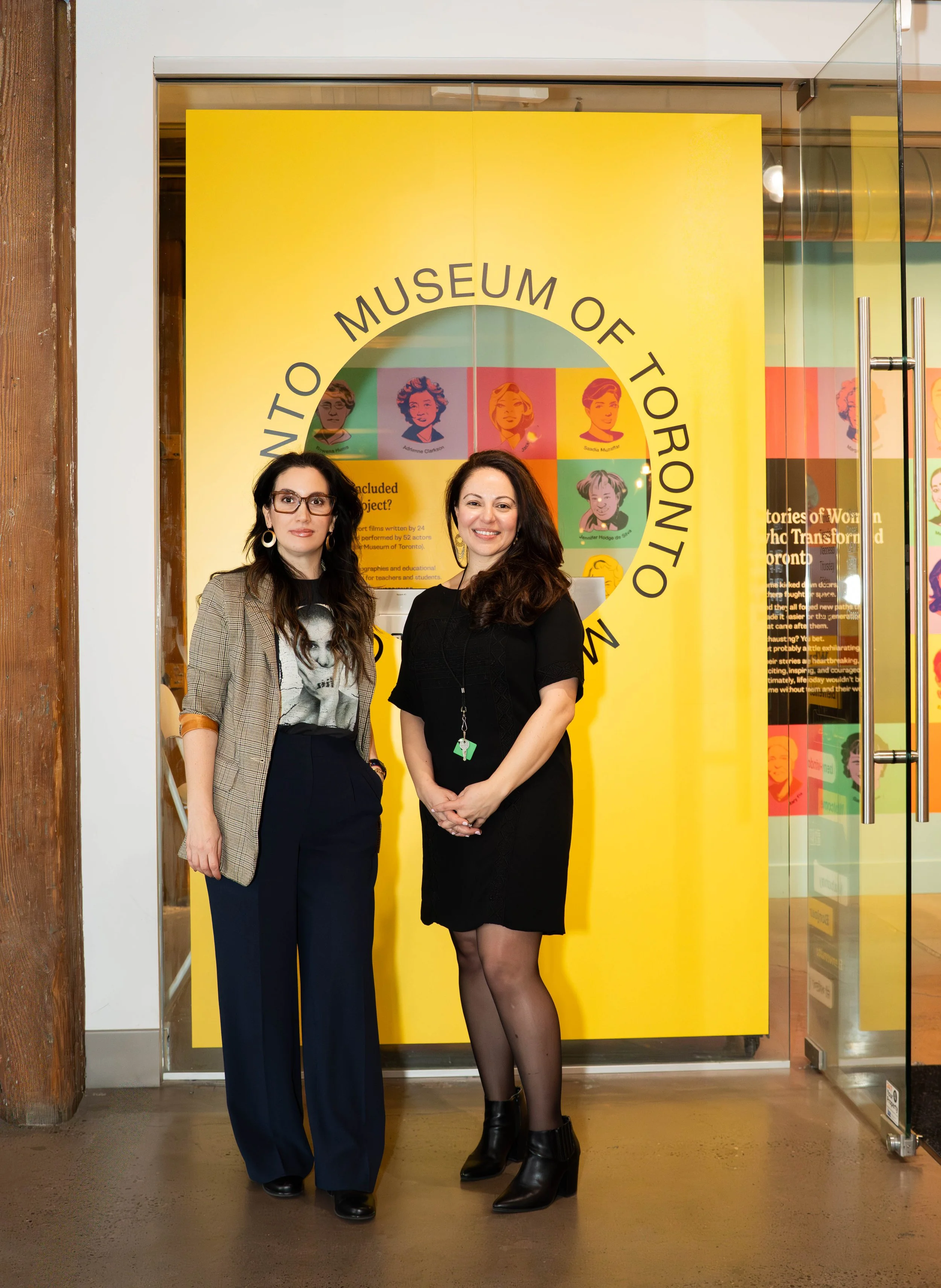 Two women standing in front of a yellow sign at the entrance of the Museum of Toronto. The woman on the left has long dark hair, glasses, and is wearing a checkered blazer over a graphic t-shirt and dark pants. The woman on the right has long dark ha