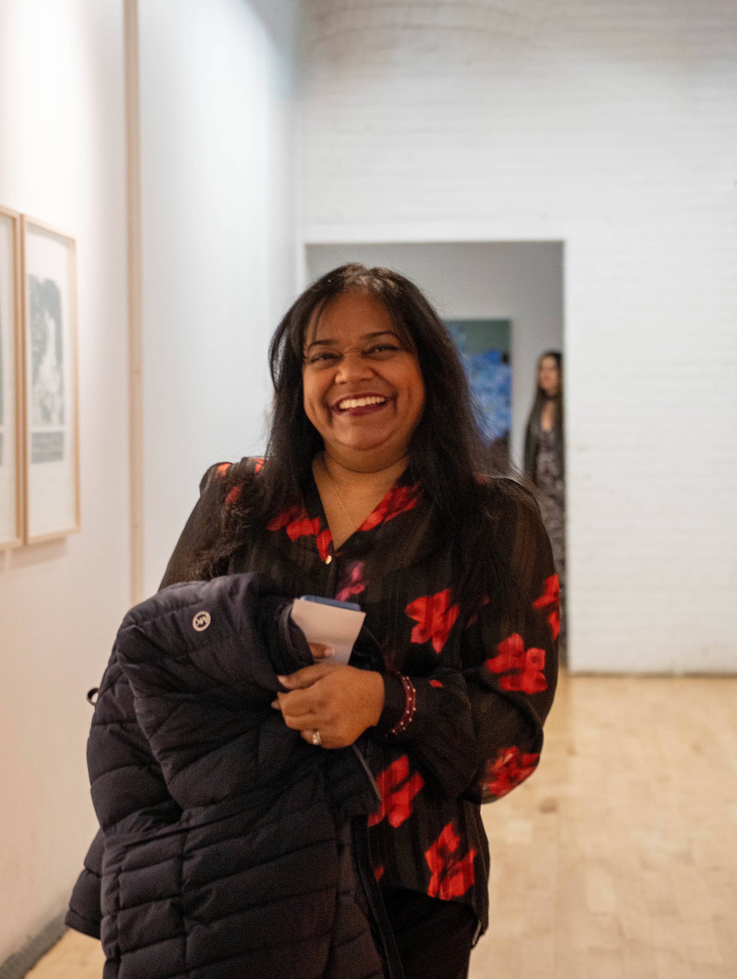 A smiling woman with dark hair and a red floral shirt holding a black puffer jacket and a notebook, standing in an art gallery hallway.