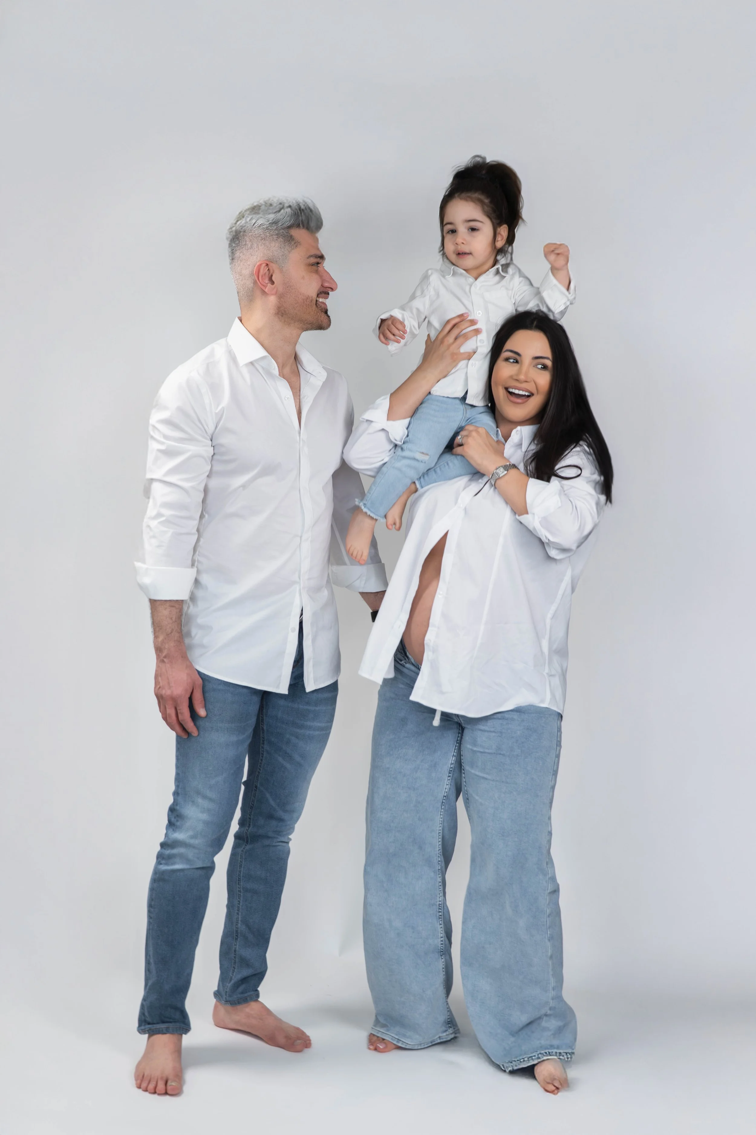 A family of three, consisting of a man, a woman, and a young girl, standing together cheerfully against a plain grey backdrop. The man and woman are barefoot and wearing white shirts and jeans, while the girl is perched on the woman’s shoulder, also 