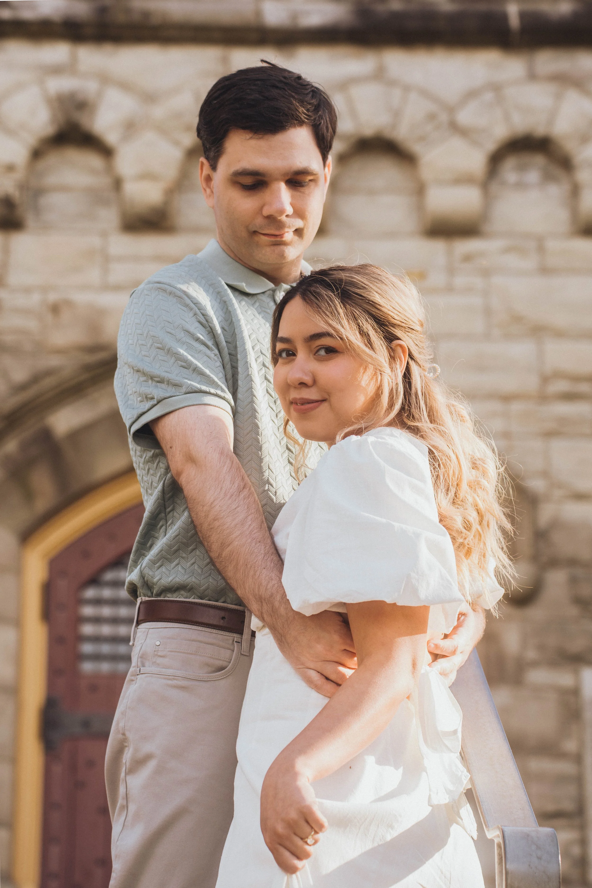 A young couple hugging outdoors in front of an old stone building with a window and arched doorway, both smiling gently.