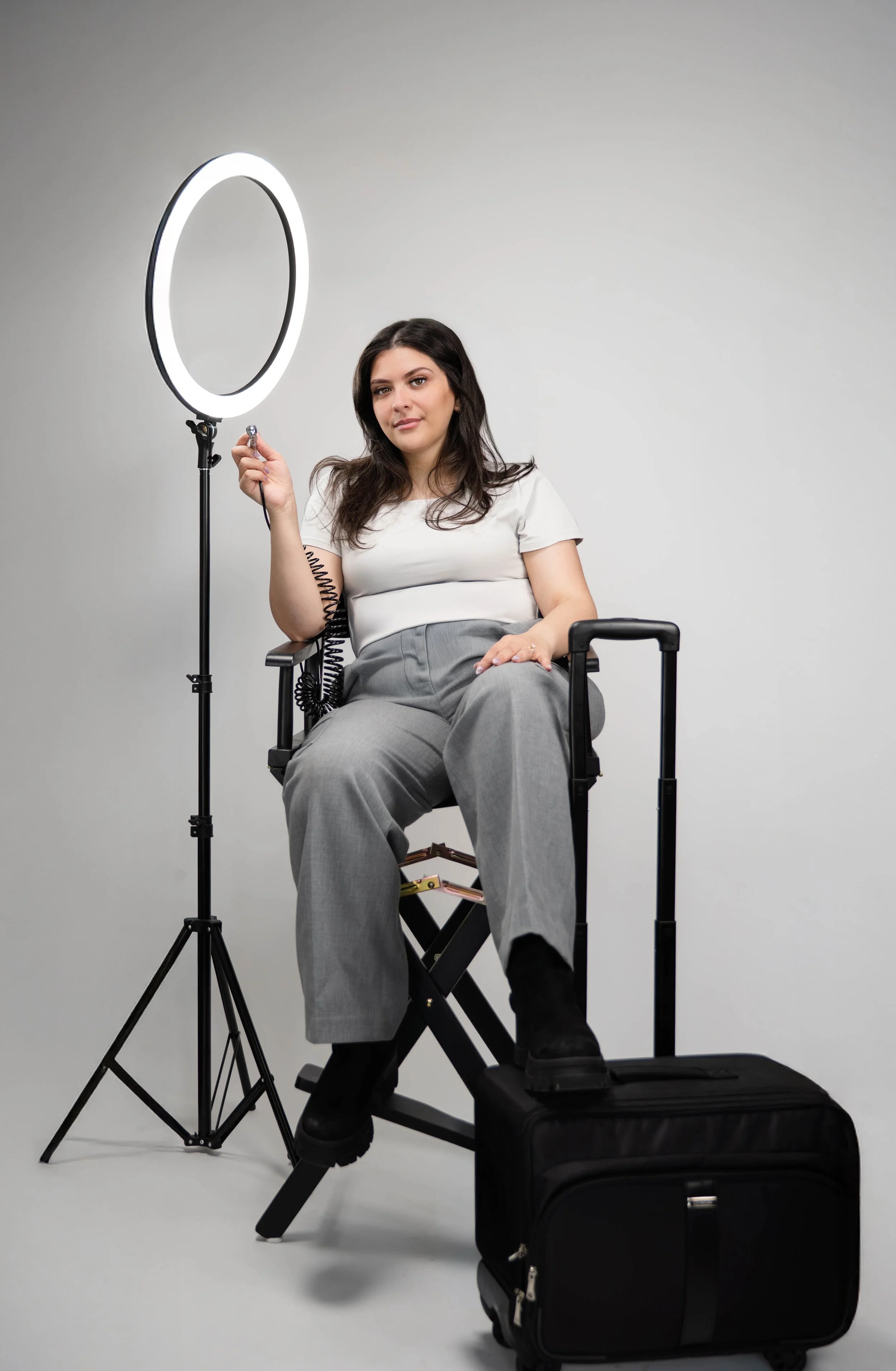 A woman with dark hair seated in a director's chair, holding a small microphone, with a ring light to her left and a rolling suitcase at her feet, against a plain gray background.
