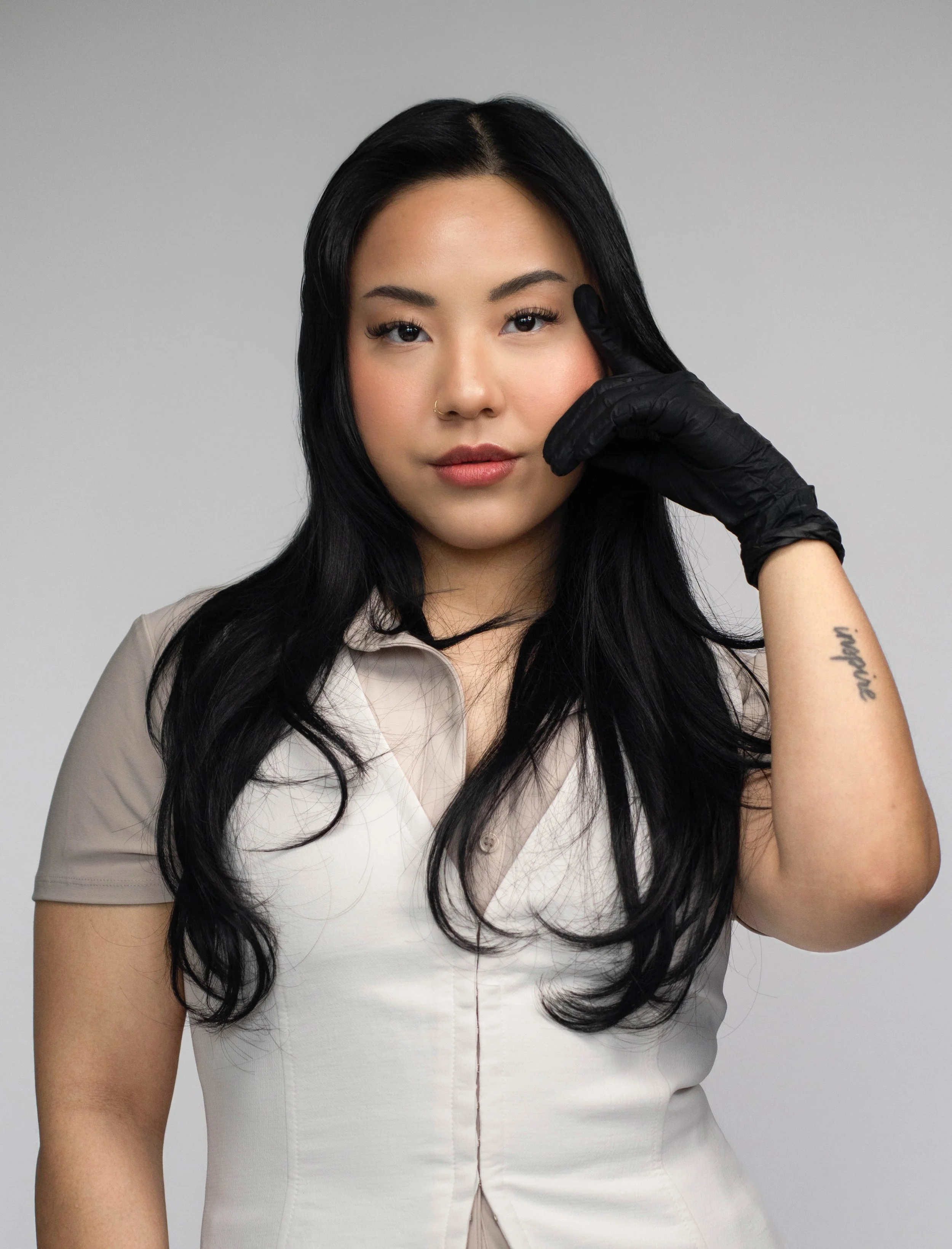 A woman with long black hair, wearing a beige and light-colored shirt, touching her face with her right hand covered by a black glove, against a neutral gray background.