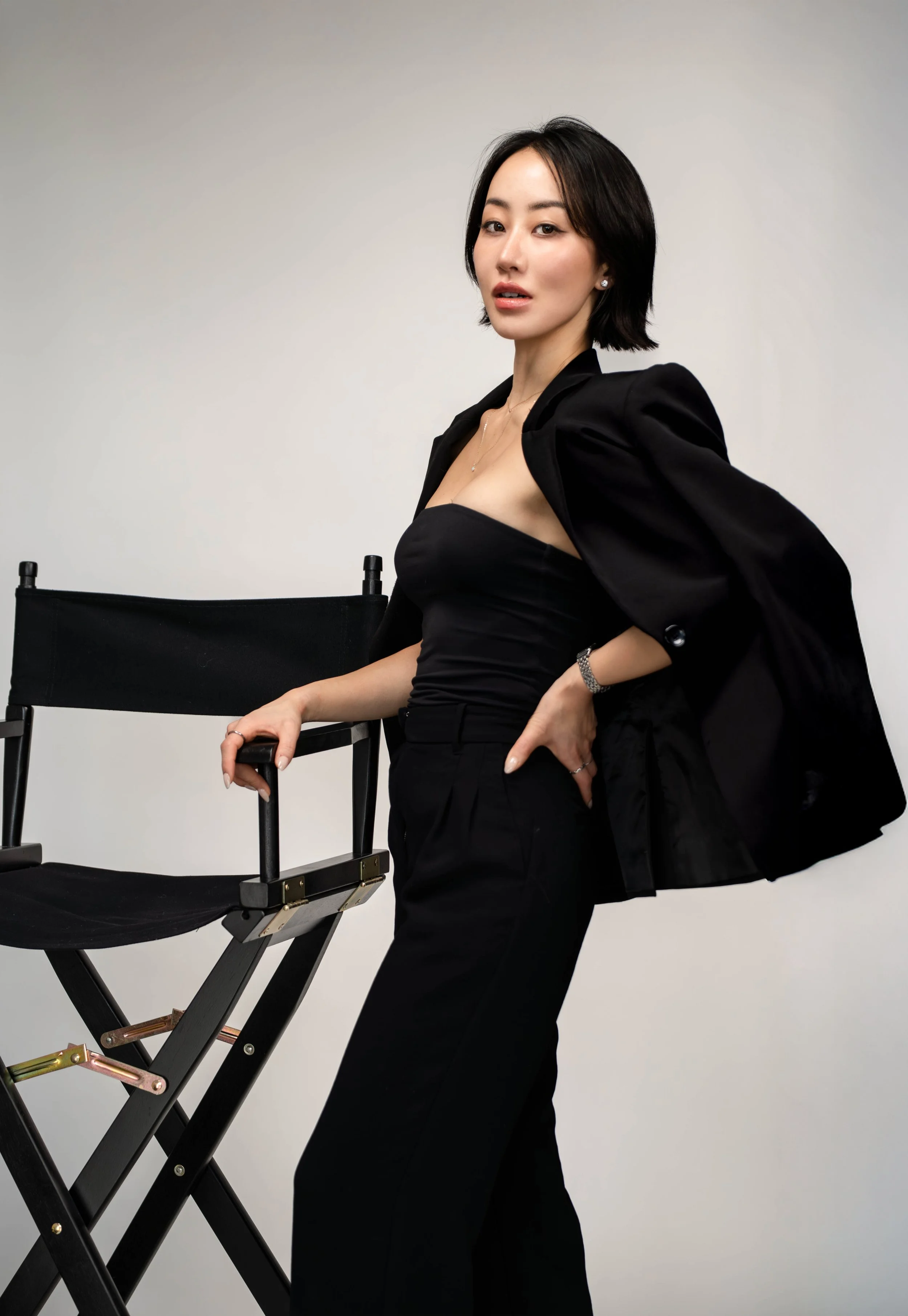 A woman with short black hair posing with a director's chair against a plain background, wearing an elegant black outfit and a black blazer draped over her shoulders.