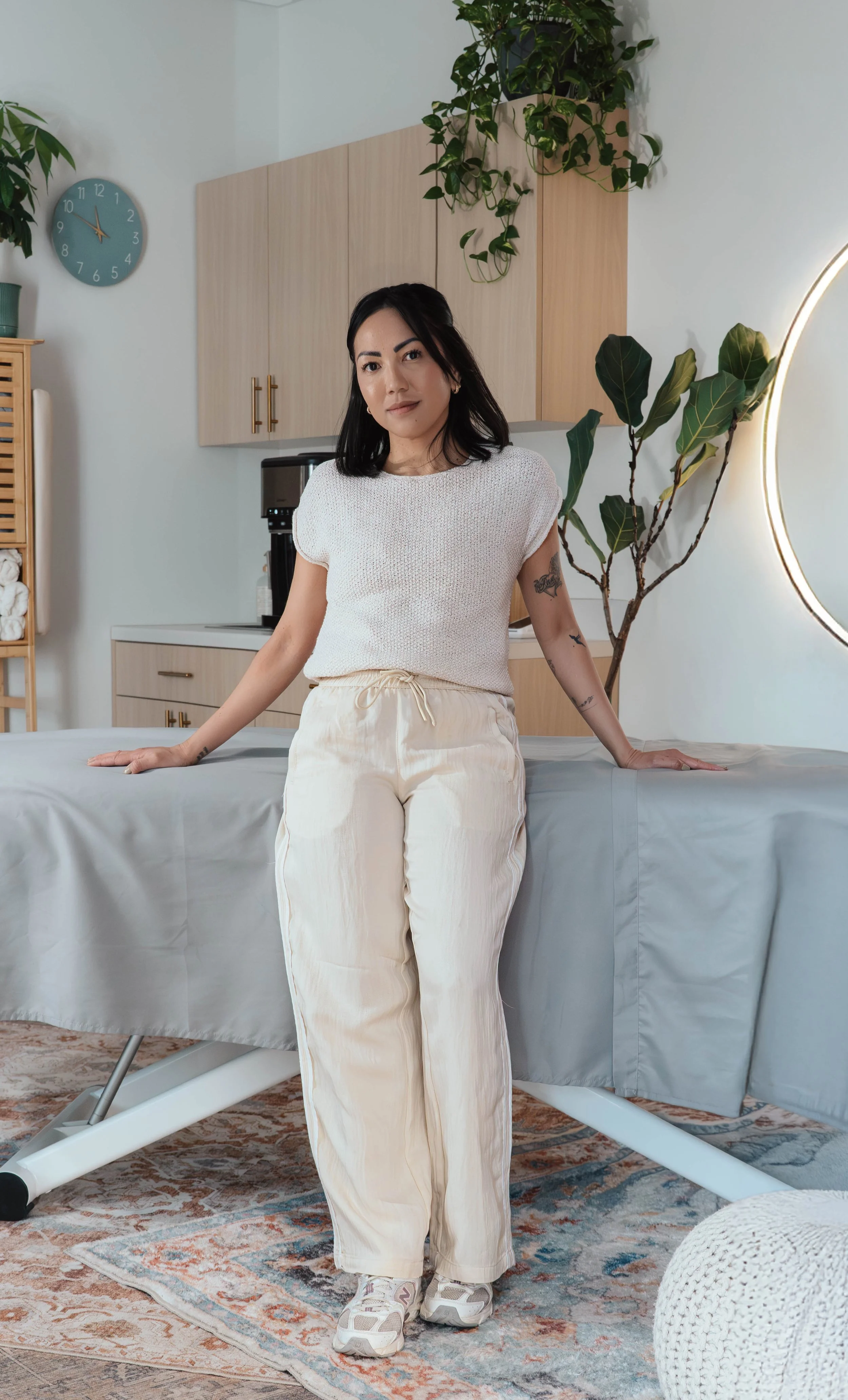 A woman with black hair, tattoos on her right arm, wearing a white knit top and cream-colored pants, sitting on a massage table in a room with wooden cabinets, green plants, a round mirror, a blue wall clock, and a patterned rug.