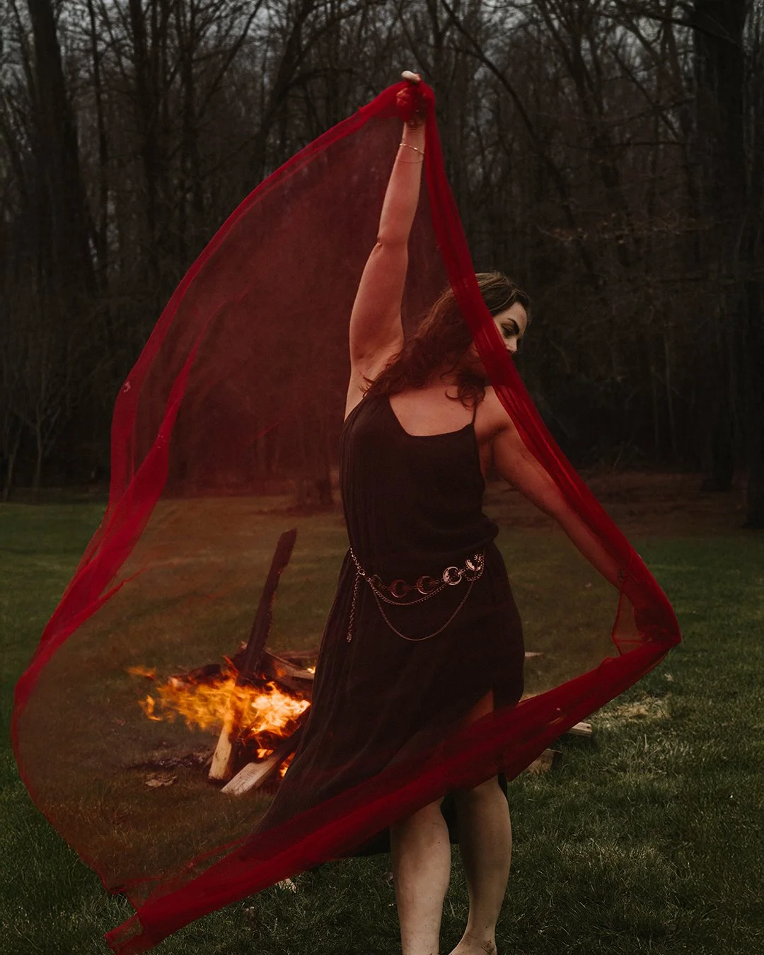 A woman standing outdoors at dusk, holding a red sheer fabric above her head. She is dressed in black and wearing silver jewelry, with a fire burning on the ground behind her.