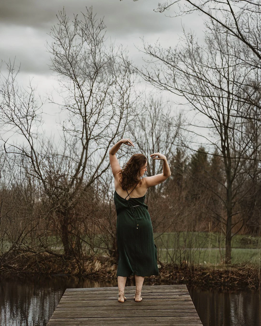 A woman with curly brown hair stands on a wooden dock by a pond, barefoot, wearing a dark green dress, with her arms raised and bent at the elbows, in a pose resembling a dancer, against a background of leafless trees and a cloudy sky.