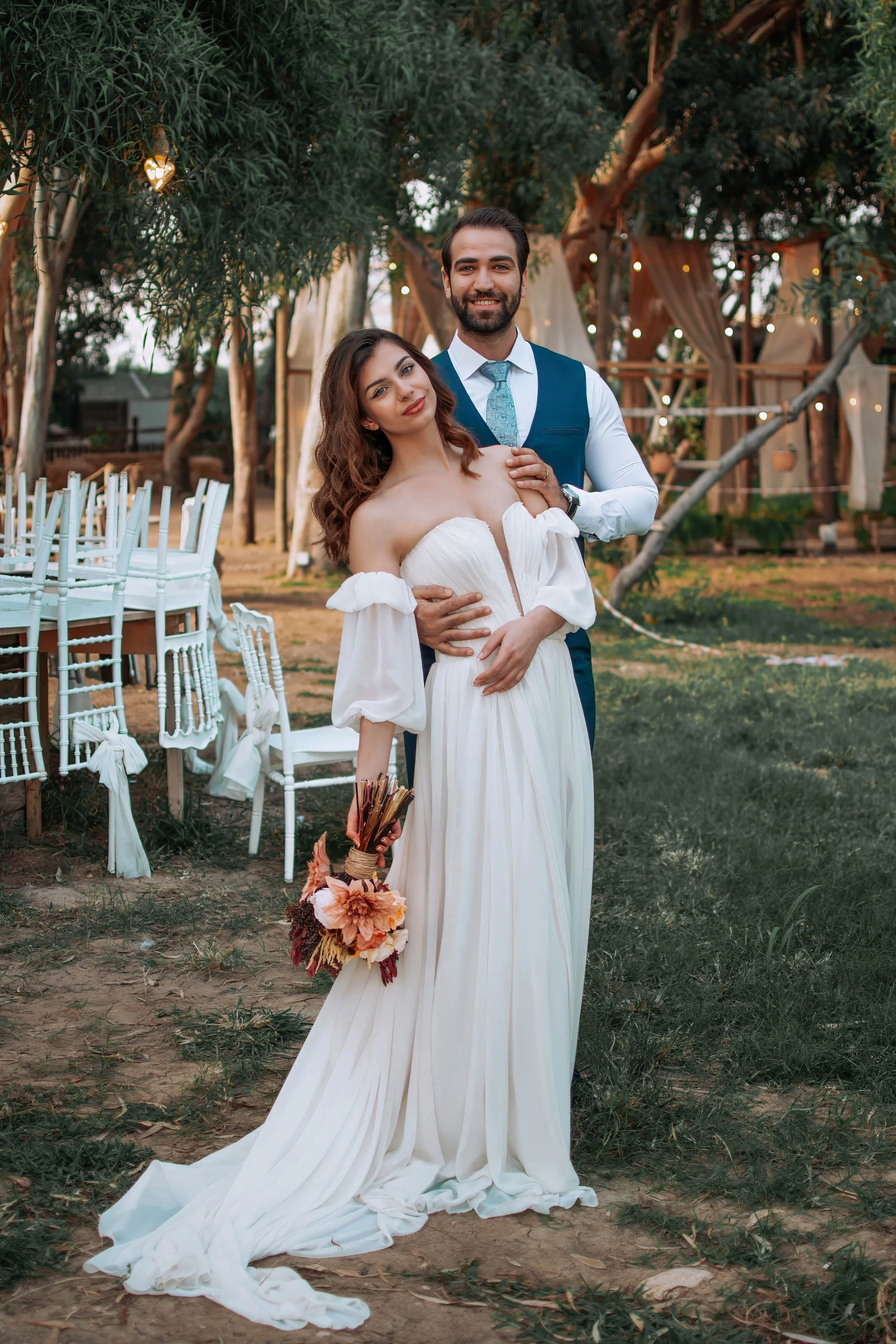 A bride and groom in wedding attire standing outdoors under trees decorated with string lights. The bride is holding a bouquet of flowers, and the groom is standing behind her with his hand on her shoulder. The scene suggests a wedding or engagement 