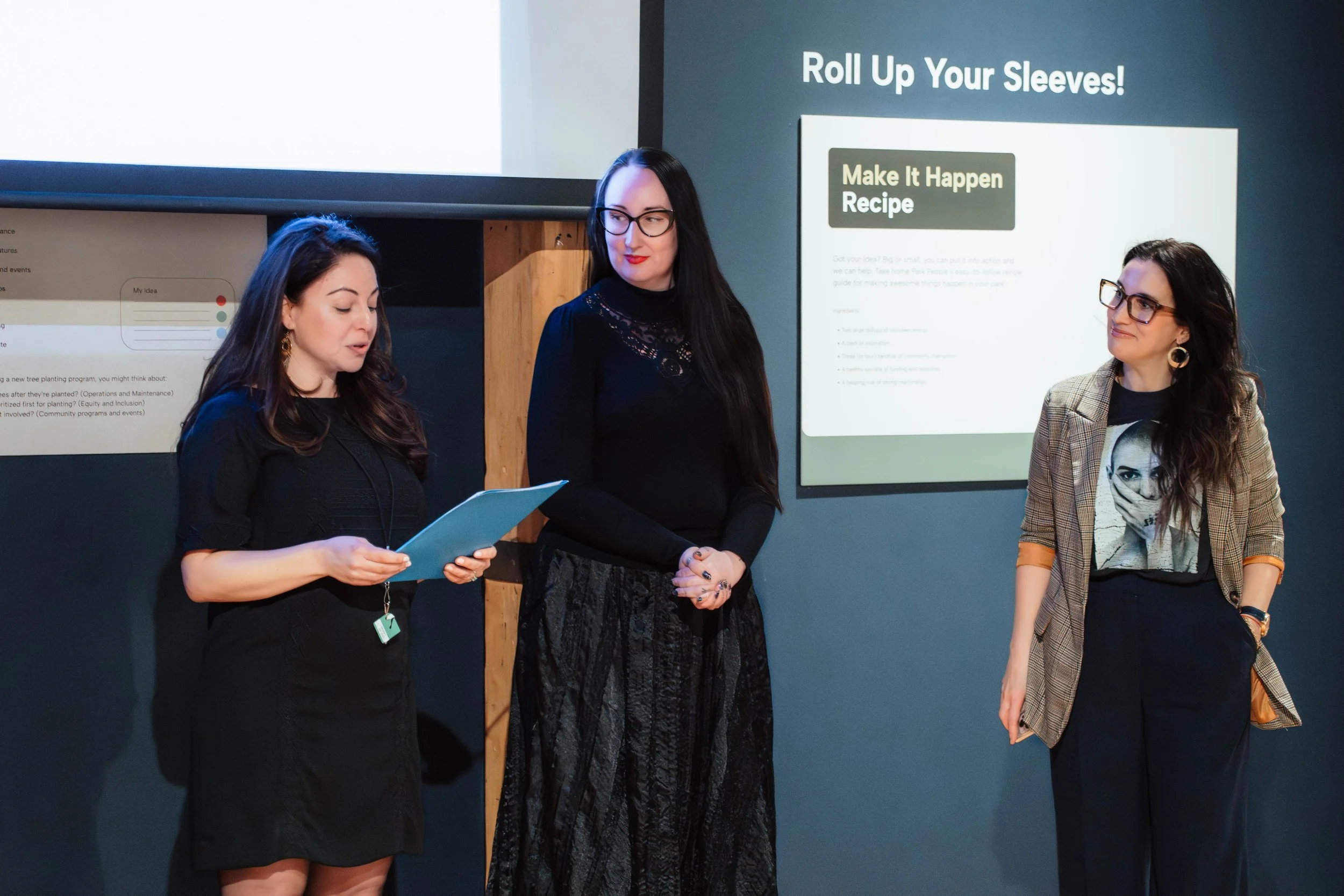 Three women standing together at a presentation. One woman is reading from a clipboard, the second woman is listening, and the third woman is looking at the speaker. Behind them are two presentation screens, one with a slide titled 'Roll Up Your Slee