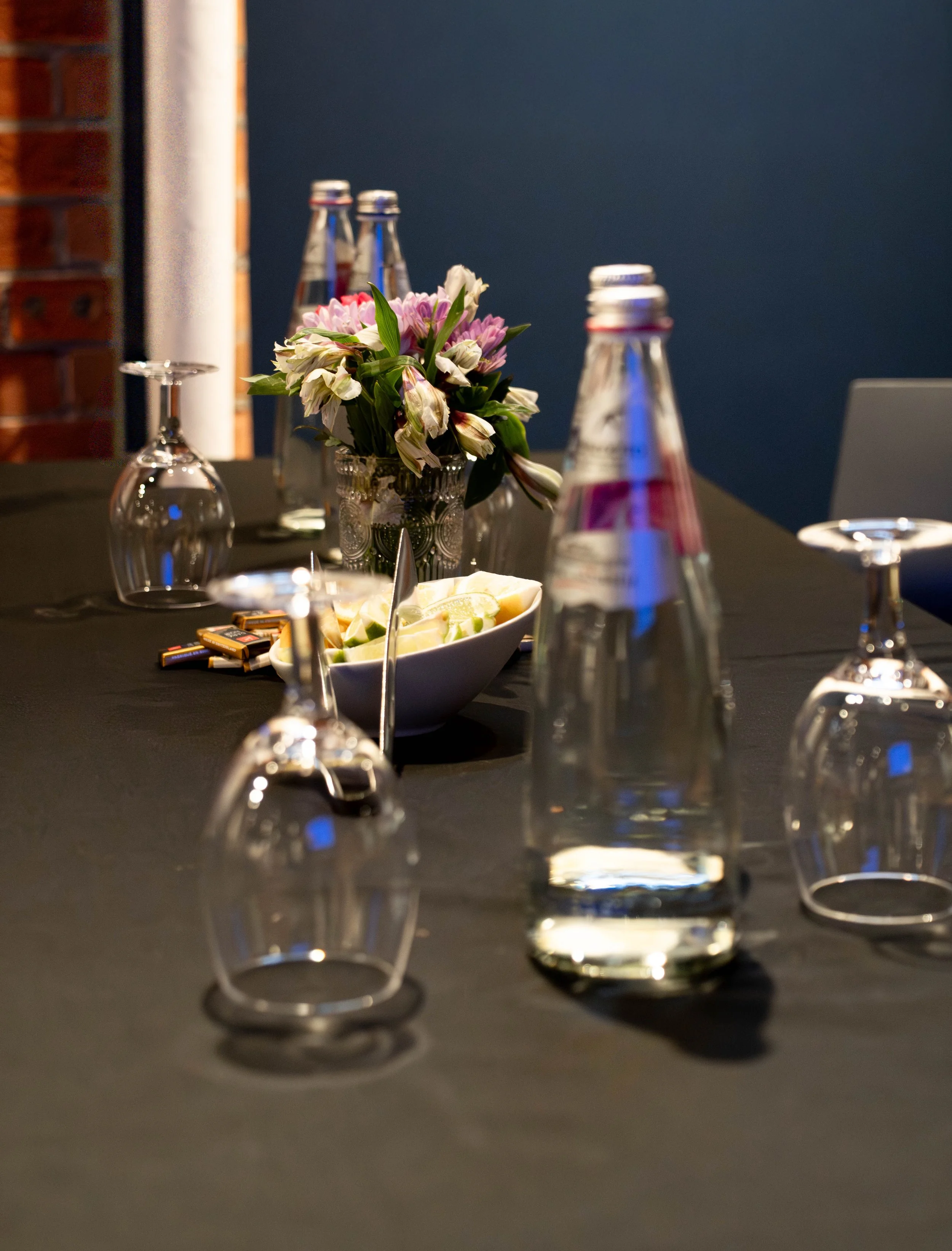A dining table set with upside-down wine glasses, a tall water bottle, a bowl of lemon wedges, a floral centerpiece with pink and white flowers, and candies, with a dark tablecloth and a blue wall background.
