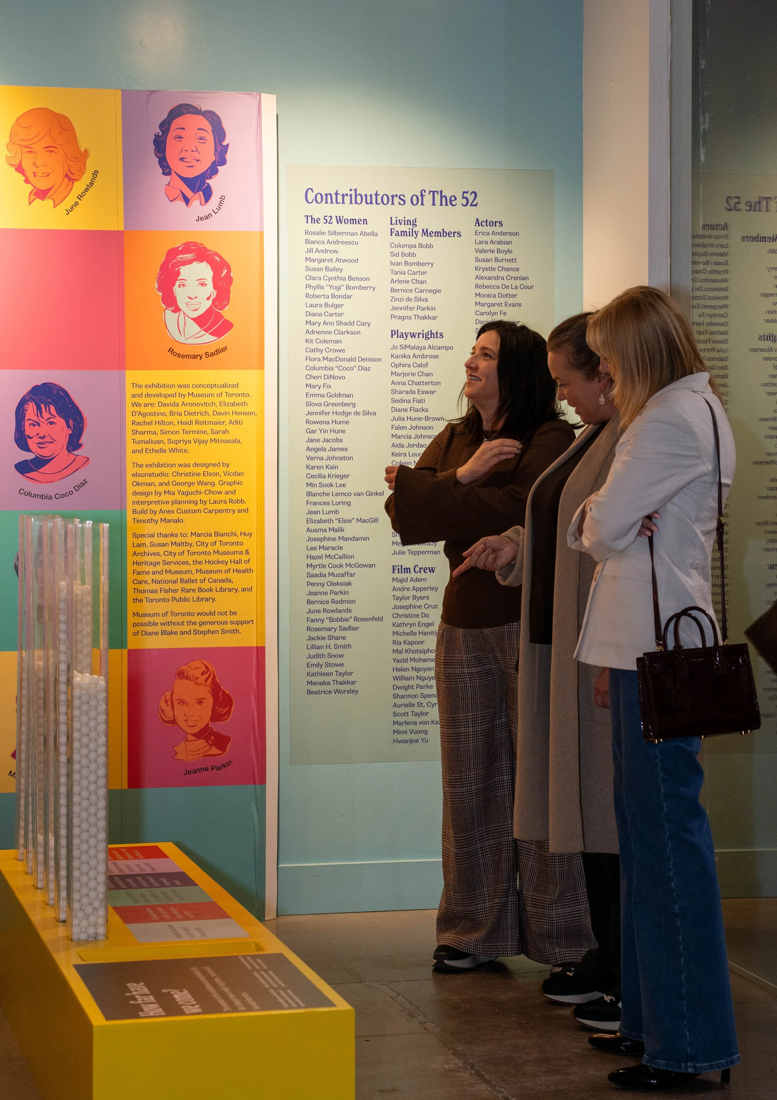 Three women stand in front of a colorful display about contributors to The 52. They are looking at the information with interest, one woman smiling and wearing a long beige coat, another smiling woman with dark hair, and a woman with blonde hair in a