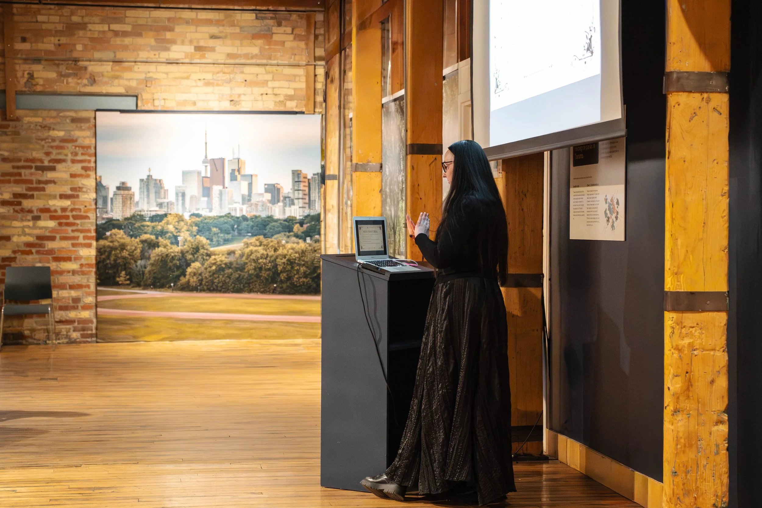 A woman with long black hair and glasses, wearing a black long-sleeve top and a long, dark patterned skirt, is gesturing with her hand while standing next to a small table with a laptop on it inside a room. Behind her is a mural of a city skyline wit