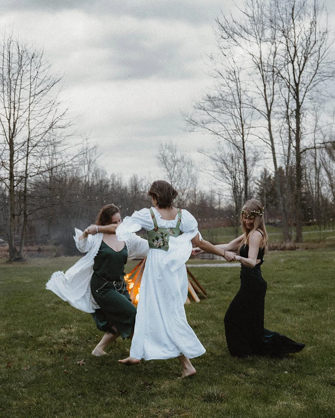 Three women dancing around a bonfire outdoors, with trees and cloudy sky in the background.