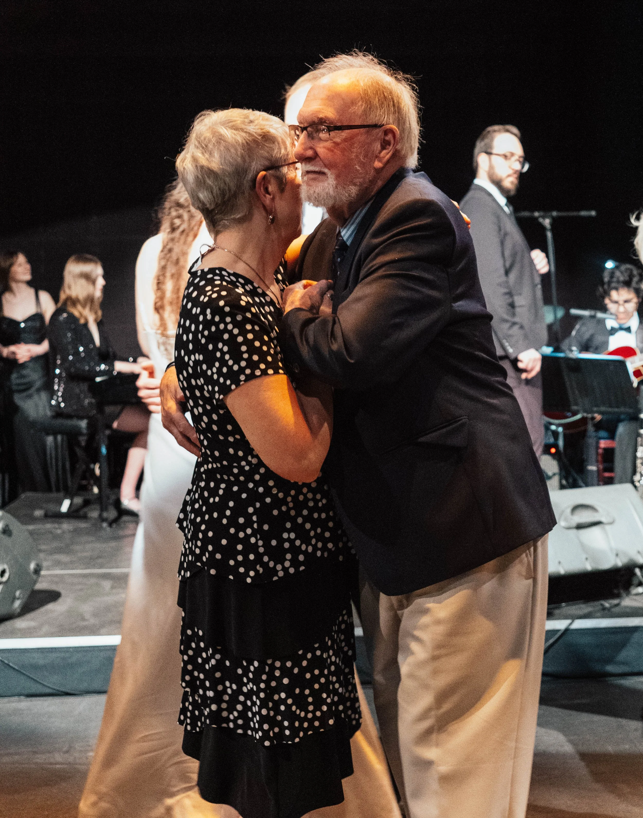 An elderly couple dancing closely together at a social event, with people and musicians in the background.