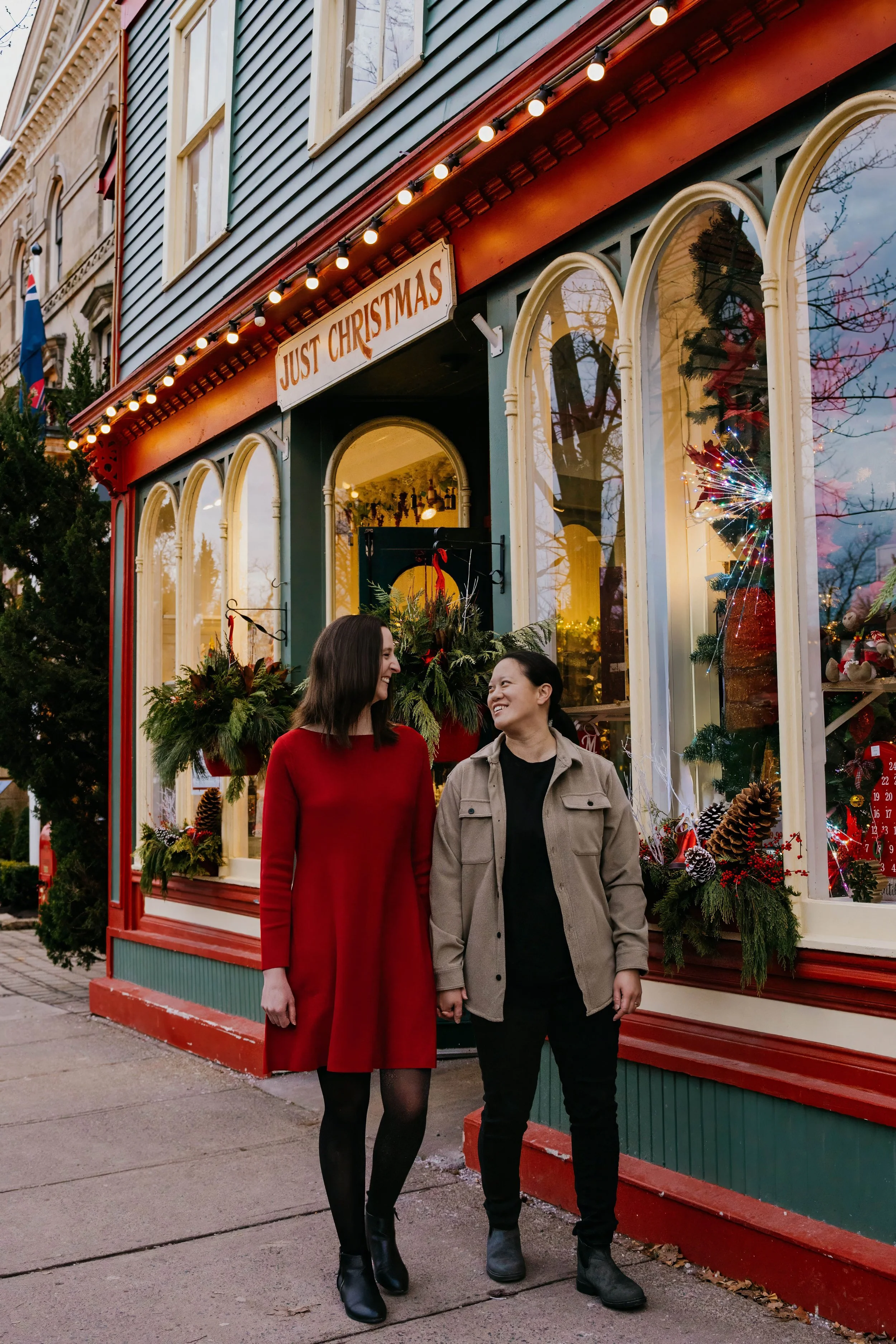Christmas couple photography in Niagara-on-the-Lake, standing together outside a festive storefront decorated with Christmas greenery, red accents and holiday decor.