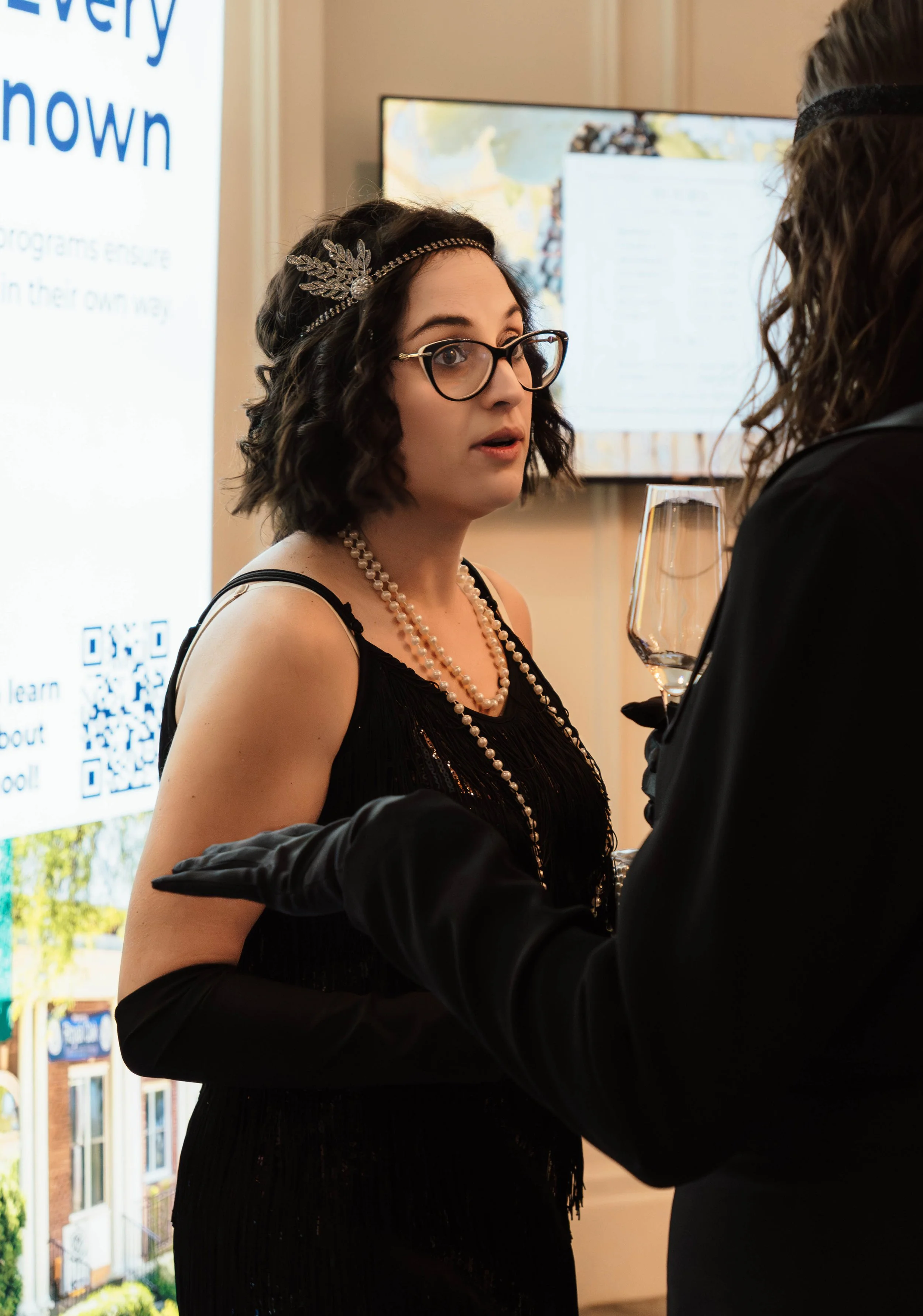 A woman with black, curly hair wearing a black dress, pearl necklace, and headband, talking to another woman holding a wine glass at an indoor event.